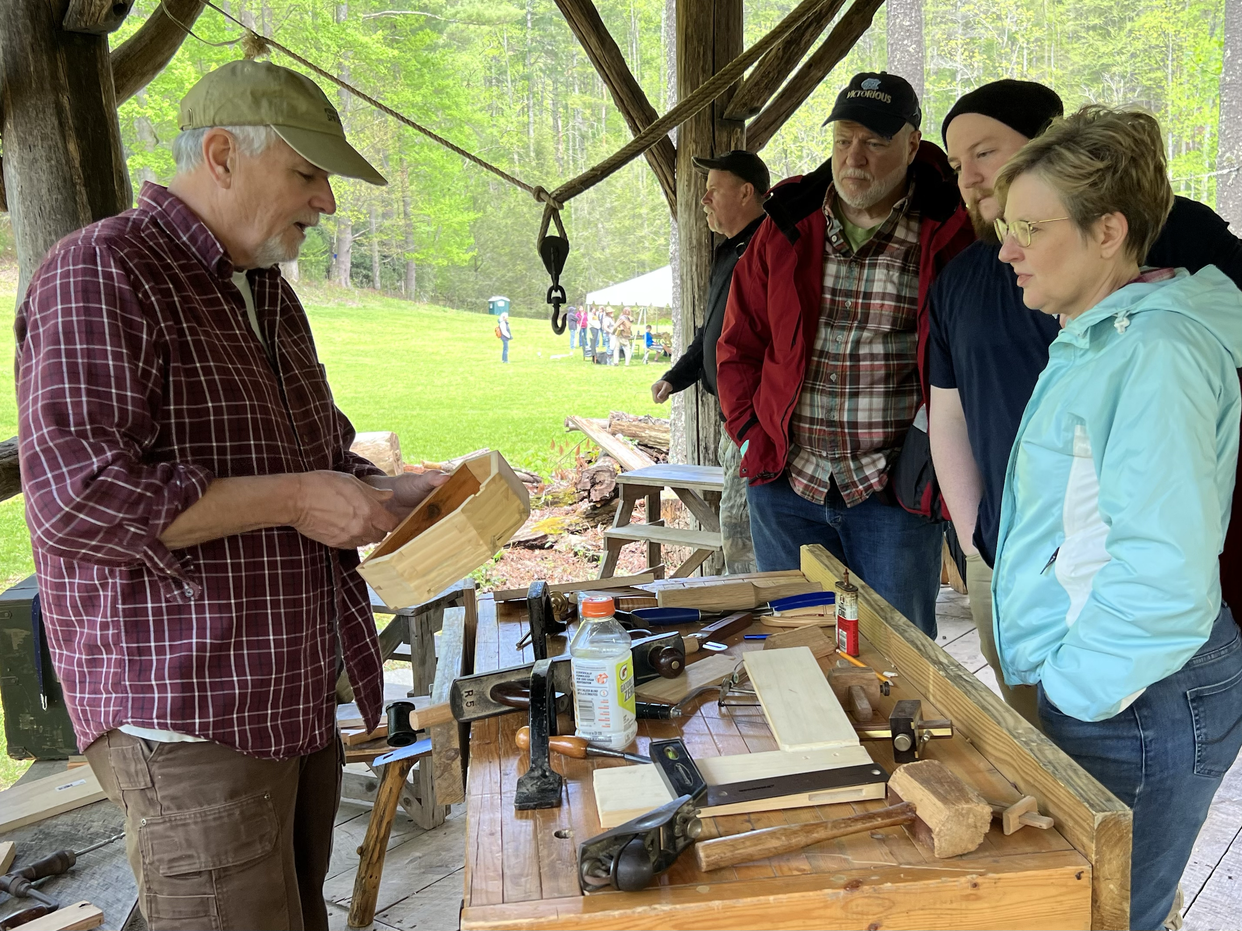 A man demonstrating woodworking to a group of four people under a wooden shelter in a park, with trees and a group of people in the background.