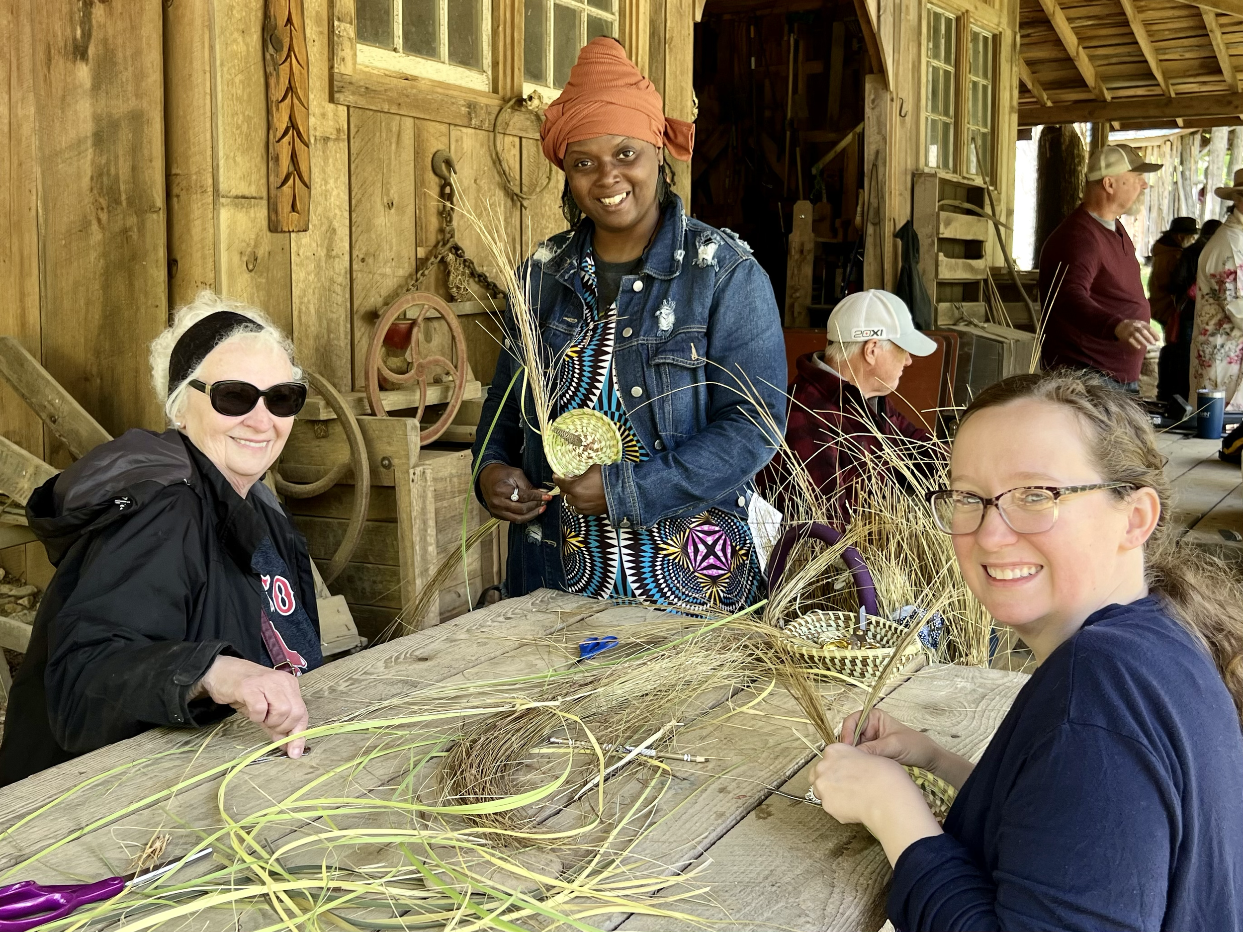 Three women sitting at a rustic wooden table outdoors, weaving straw or grass. One woman with sunglasses and dark clothing is smiling, another woman with glasses is also smiling, and a third woman with a headscarf is standing and holding some woven i