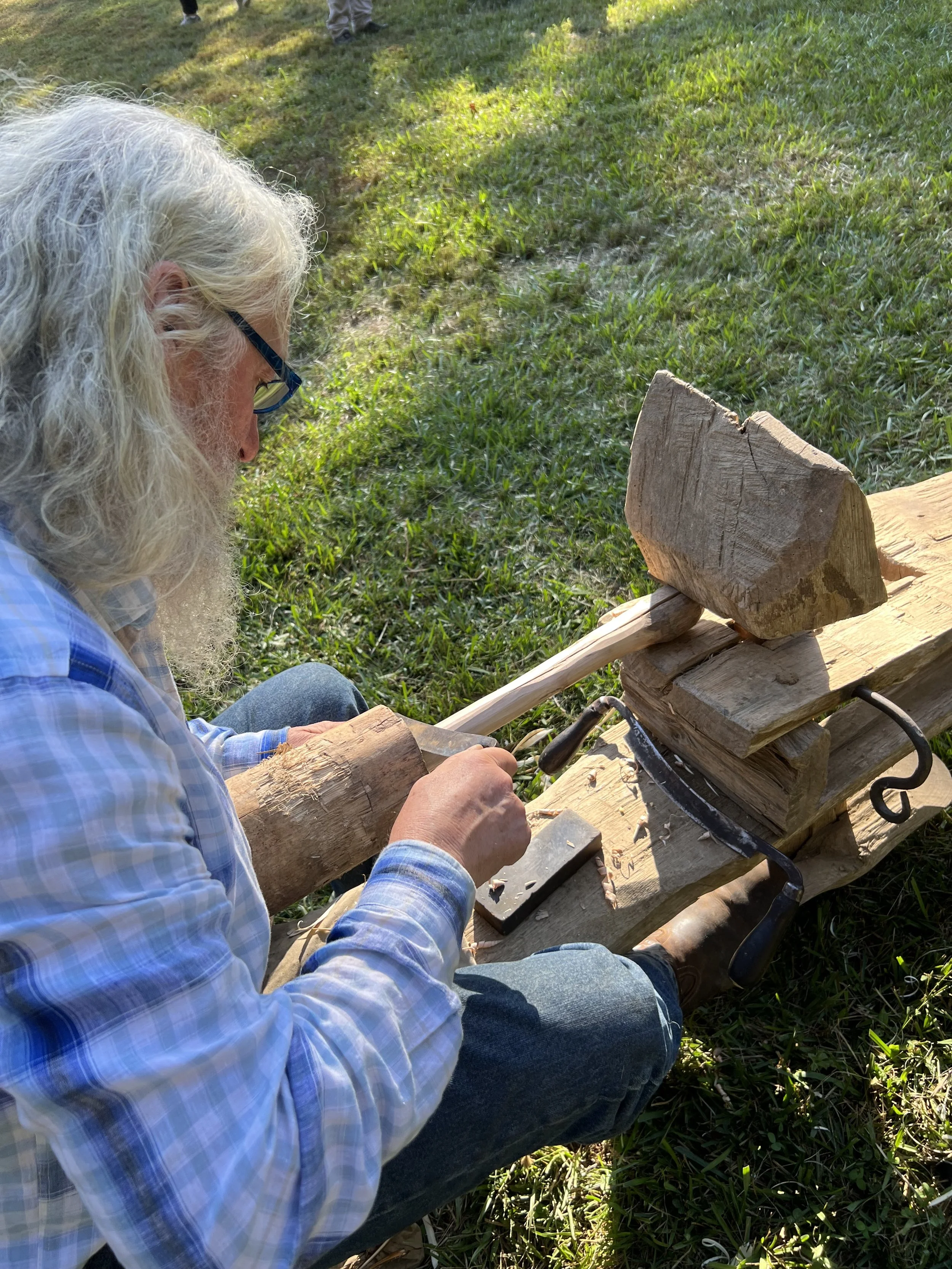 An elderly man with long white hair and glasses, wearing a blue plaid shirt and jeans, is sitting outside on grass, carving a large wooden log using a chisel and hammer. He is focused on his woodworking project, with wooden pieces and tools around him.