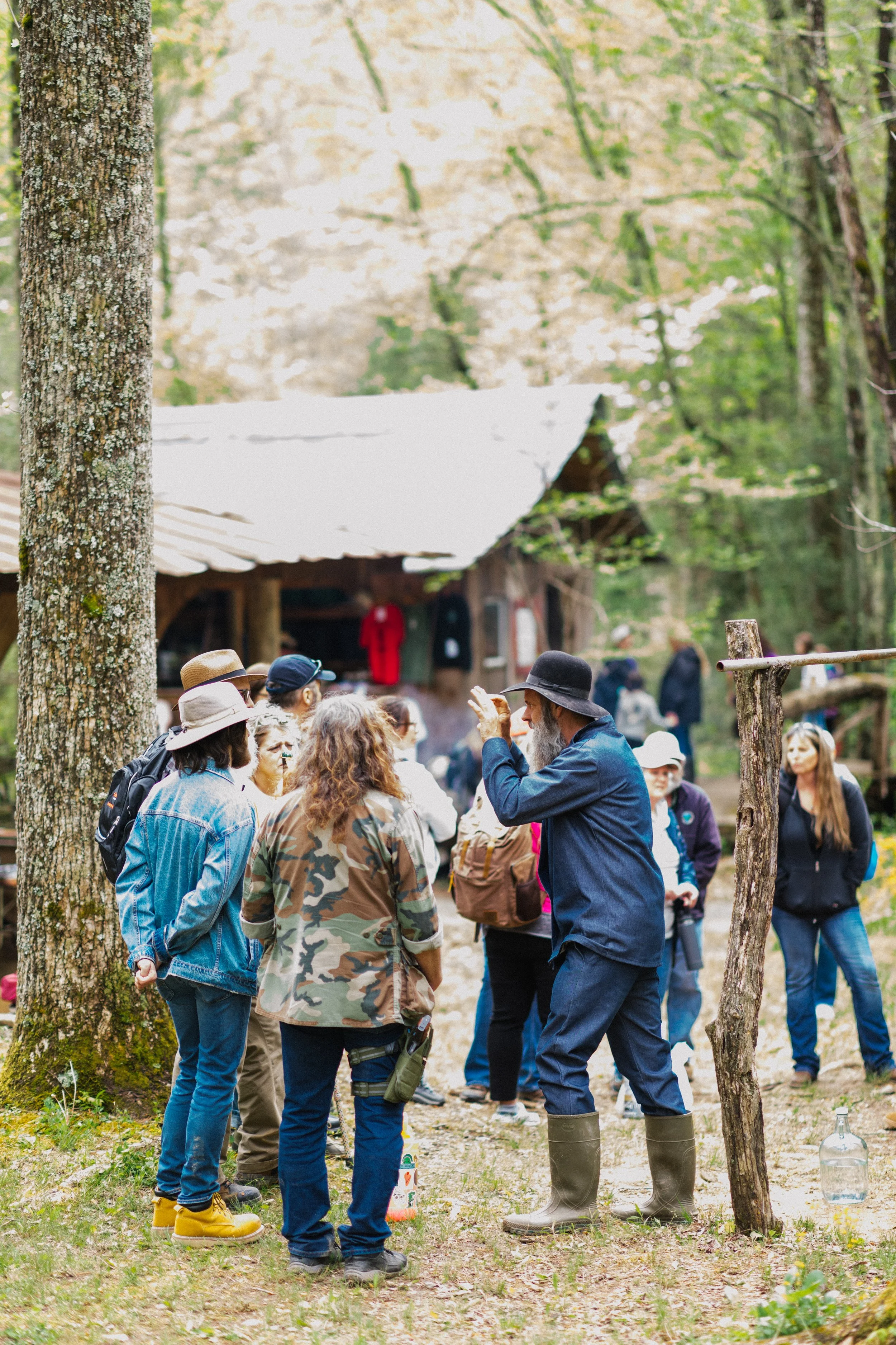 A group of people gathered outdoors near a rustic wooden cabin in a forest, listening to a man in a black hat and boots explain something.