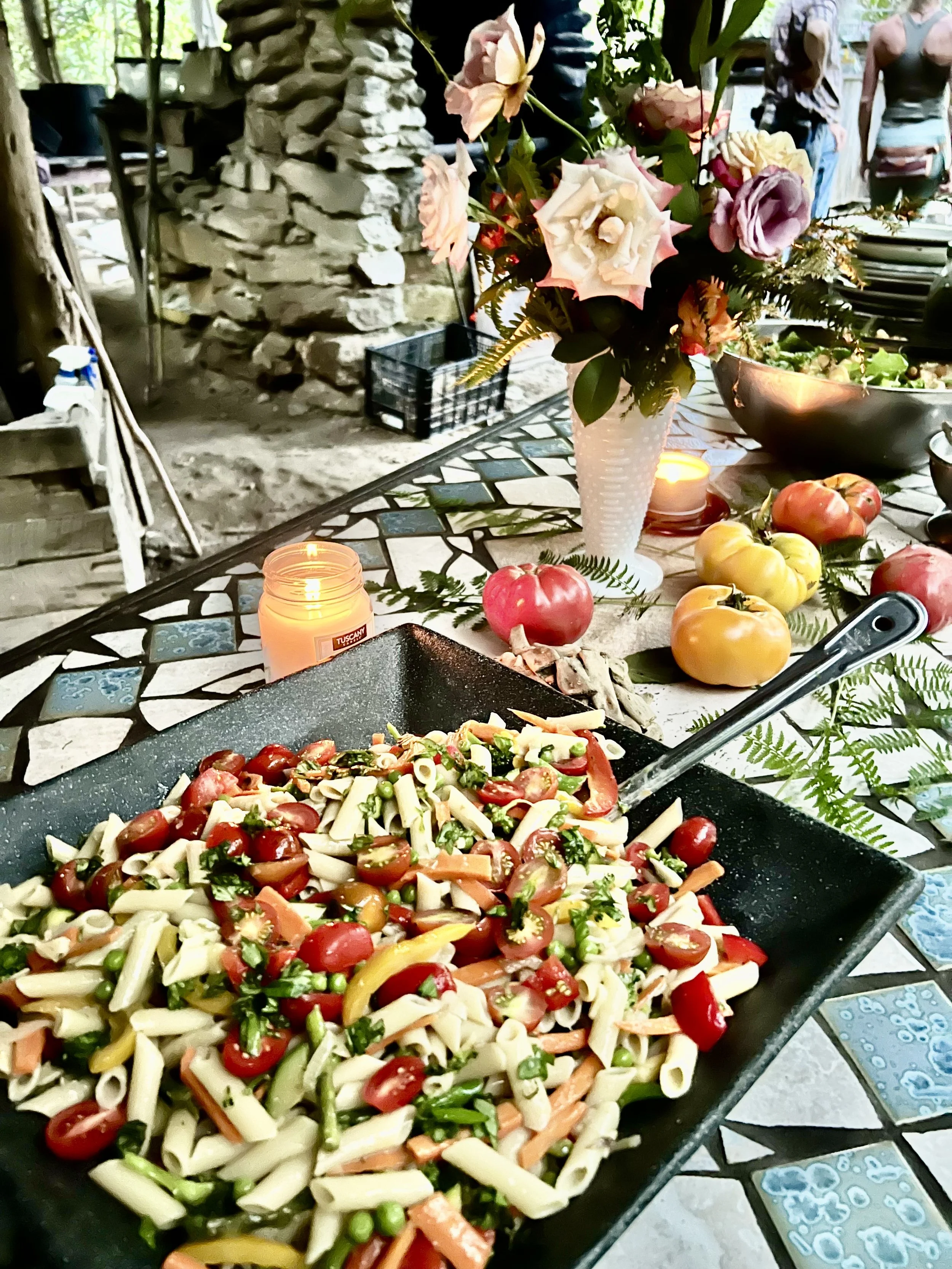 A skillet of pasta salad with cherry tomatoes, green peas, and chopped vegetables on a mosaic table, surrounded by fruit, flowers, and candles at an outdoor gathering.