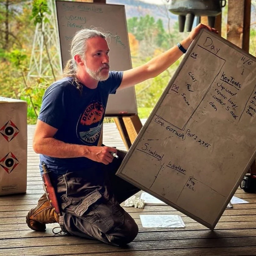 Man with long gray hair and beard, wearing a blue t-shirt and camouflage cargo pants, kneeling on wooden deck, holding a whiteboard with notes about kayaking, talking to an audience.