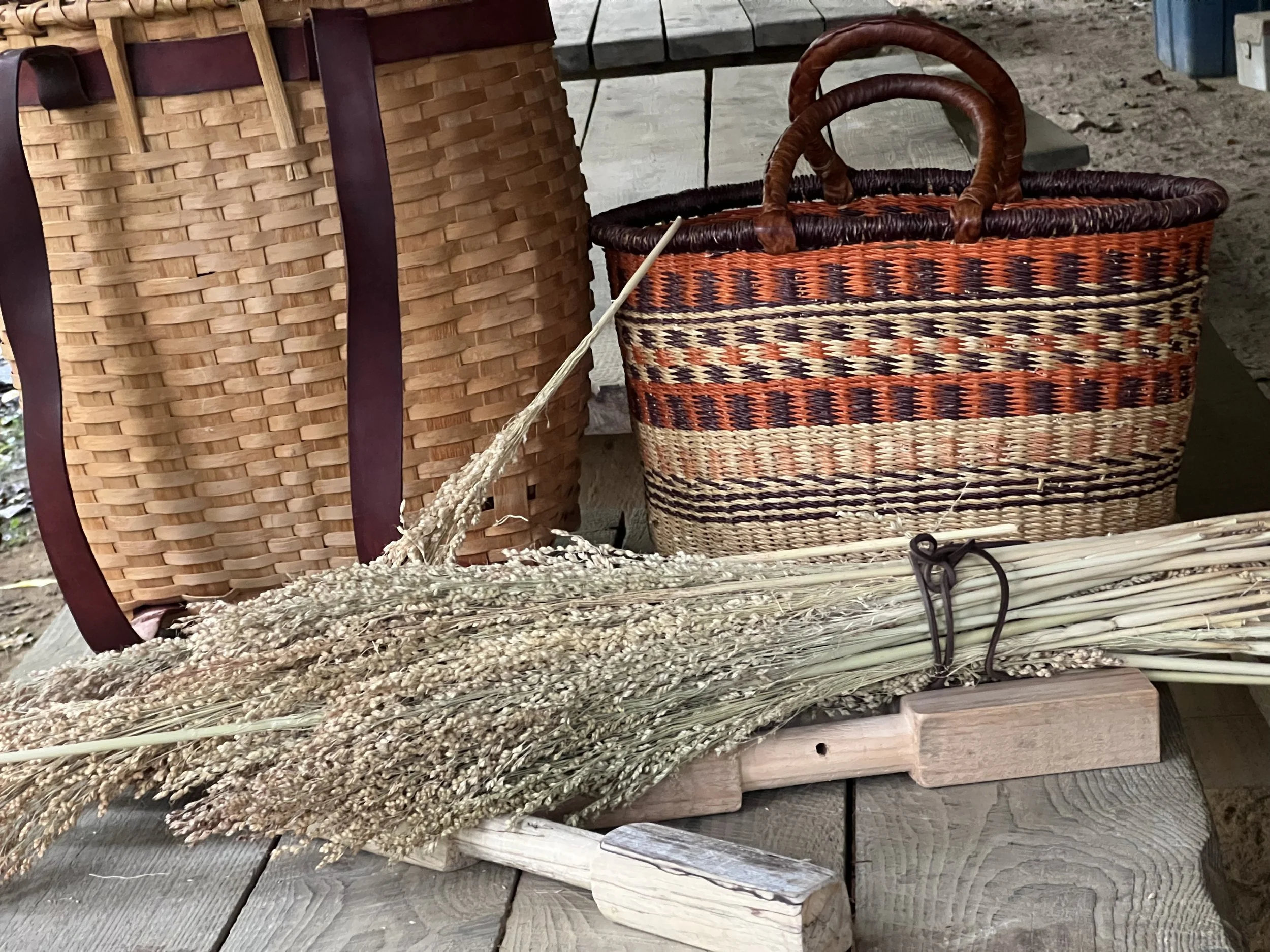 Bunch of dried wheat stalks lying on a wooden surface in front of two woven baskets, one tall and one short, on a rustic table.
