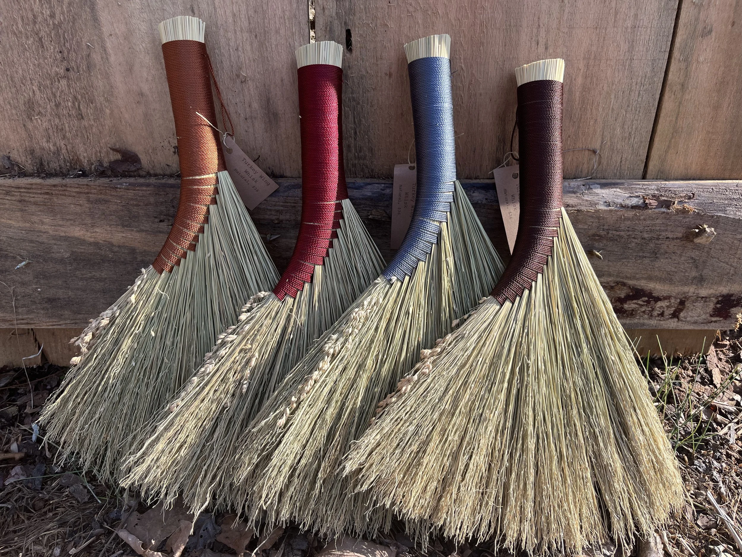 Four handmade brooms with different colored handles (red, pink, blue, brown) leaning against a wooden surface outdoors with soil and leaves at the base.