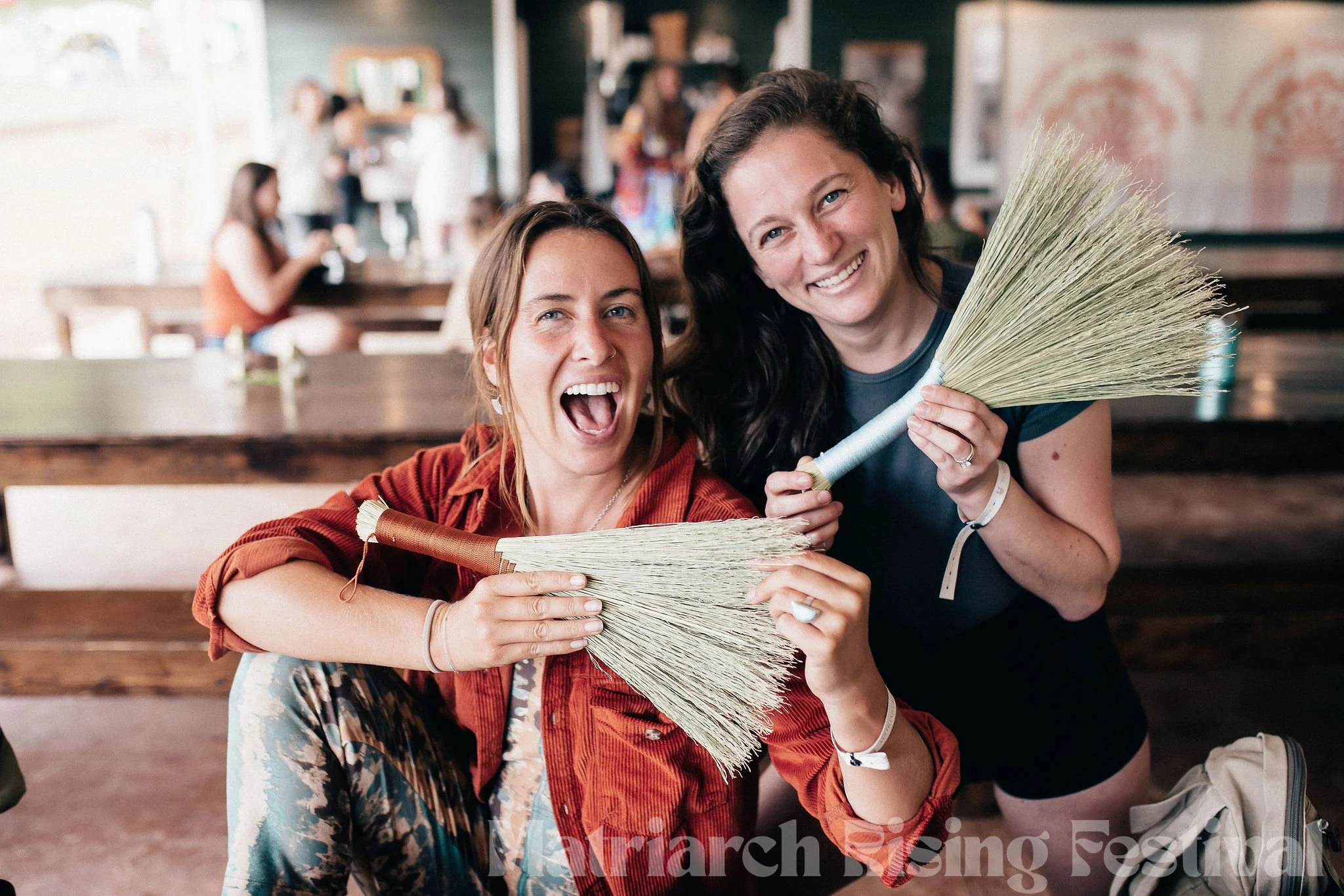 Two women smiling and holding brooms at a festival with other people sitting at tables in the background.