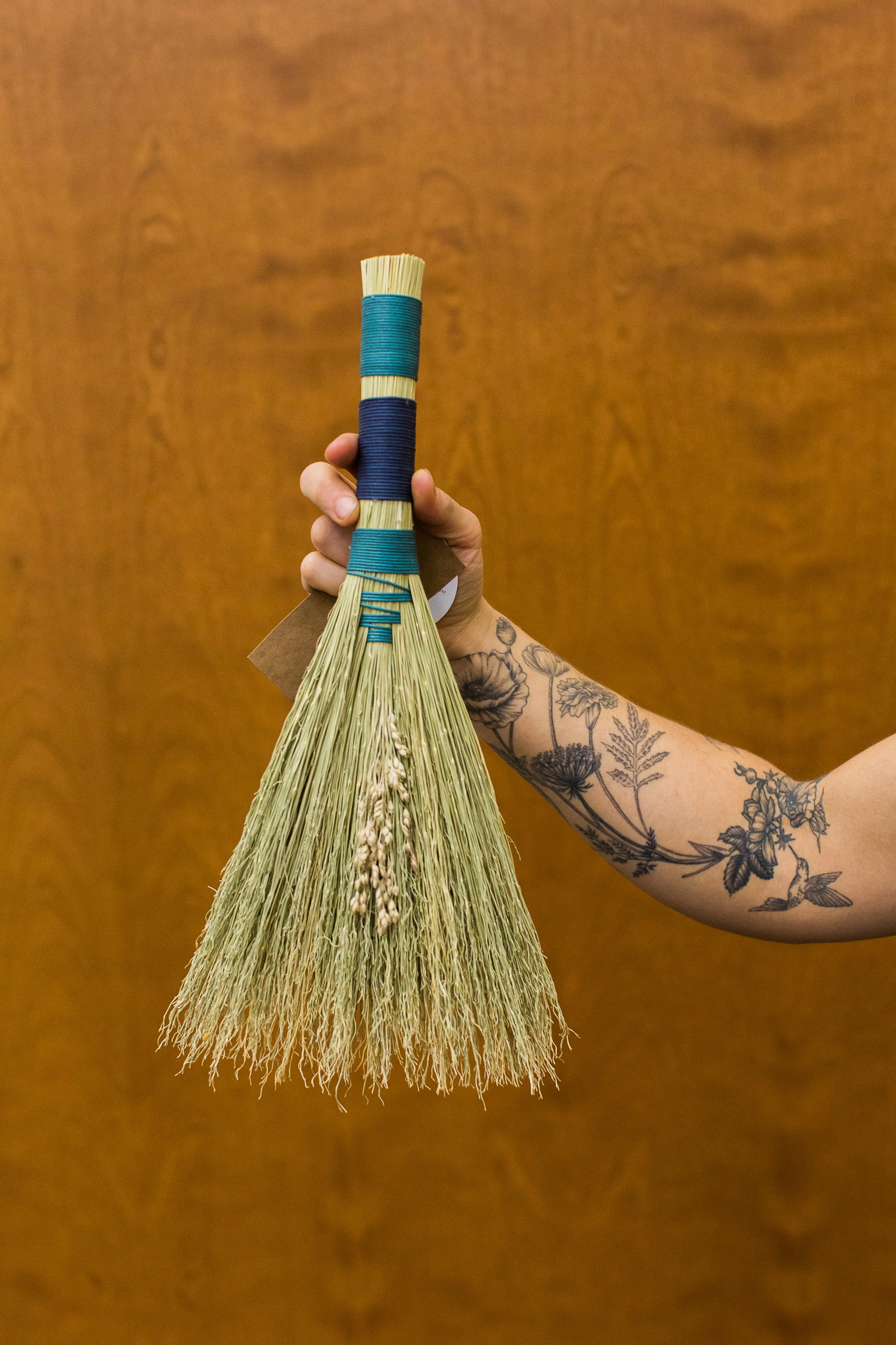 A hand with floral tattoos holding a decorative broom made of wheat stalks with a handle wrapped in teal and navy thread, against a wooden background.