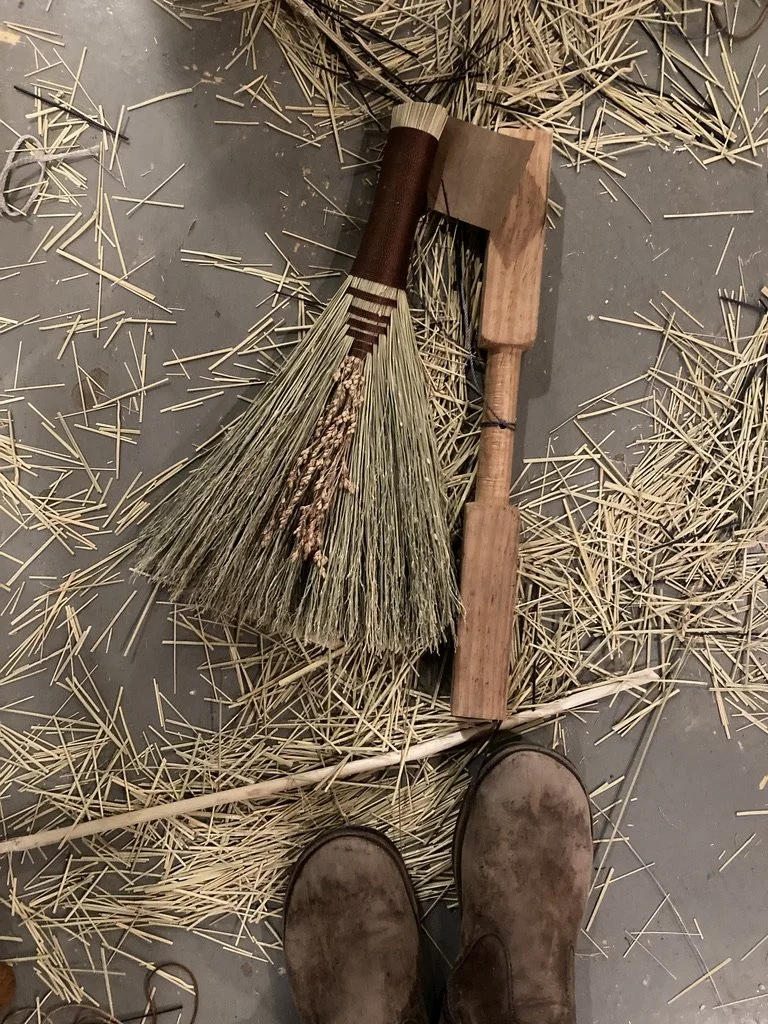 A broom and a small axe lying on a concrete floor surrounded by scattered straw and hay, with a person's worn boots visible at the bottom of the image.