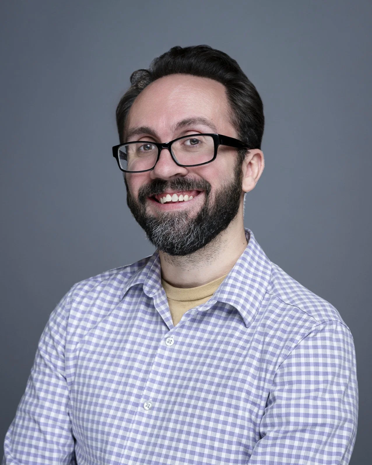 Headshot of a smiling man with glasses, a beard, and dark hair, wearing a checkered shirt, against a gray background.