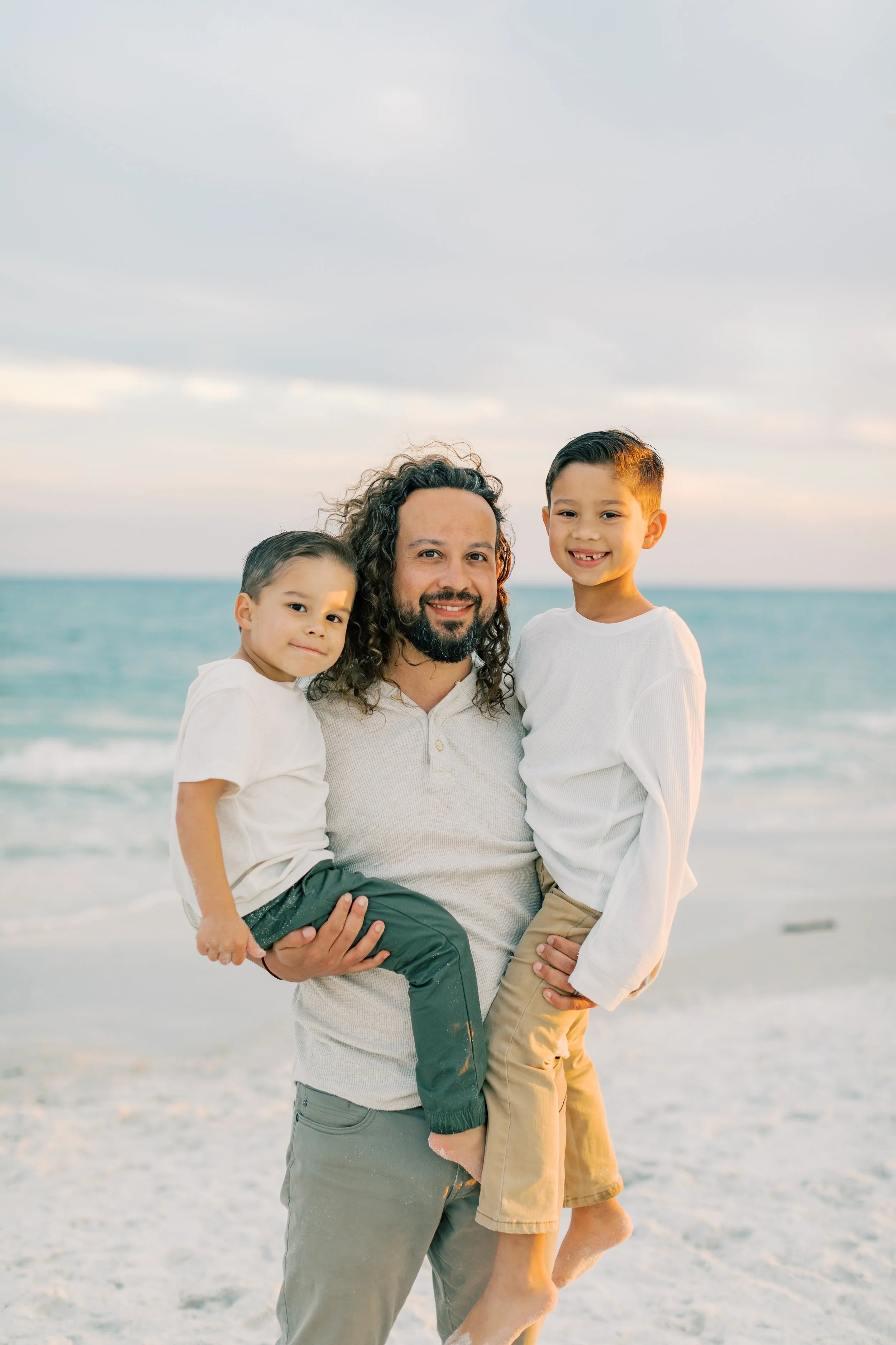 A man with curly hair and a beard holding two children, standing on a beach with the ocean and sky in the background.