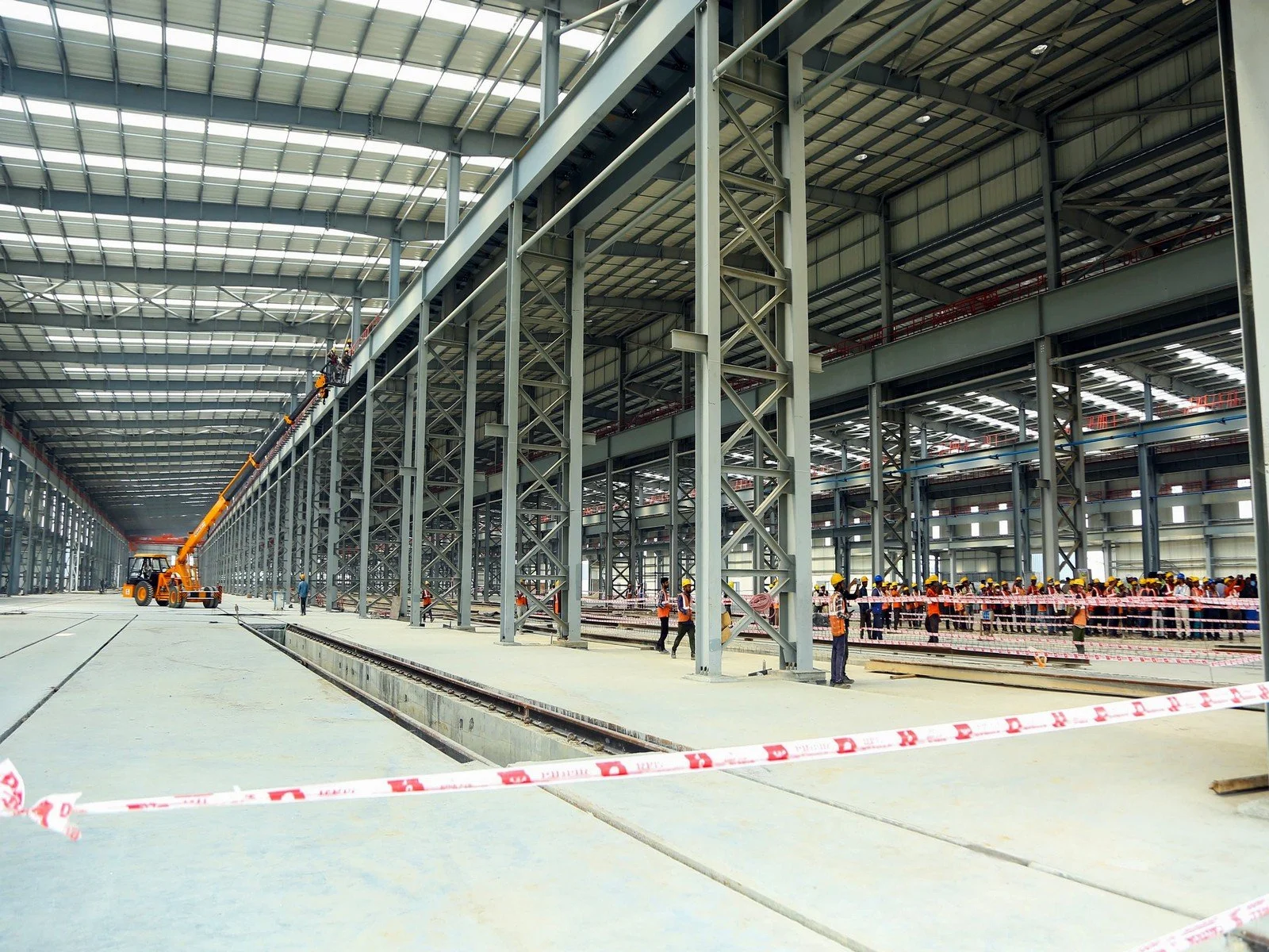 Construction site inside a large industrial building with steel framework, workers wearing safety helmets and vests, and a crane lifting materials.