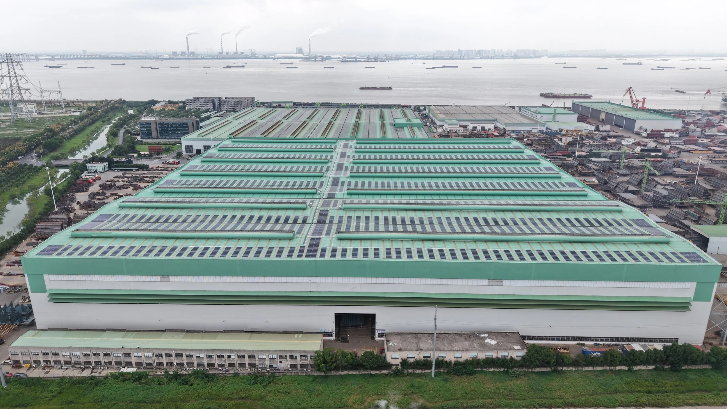 An industrial building with solar panels on the roof, located near a river with ships, and a cityscape in the background.