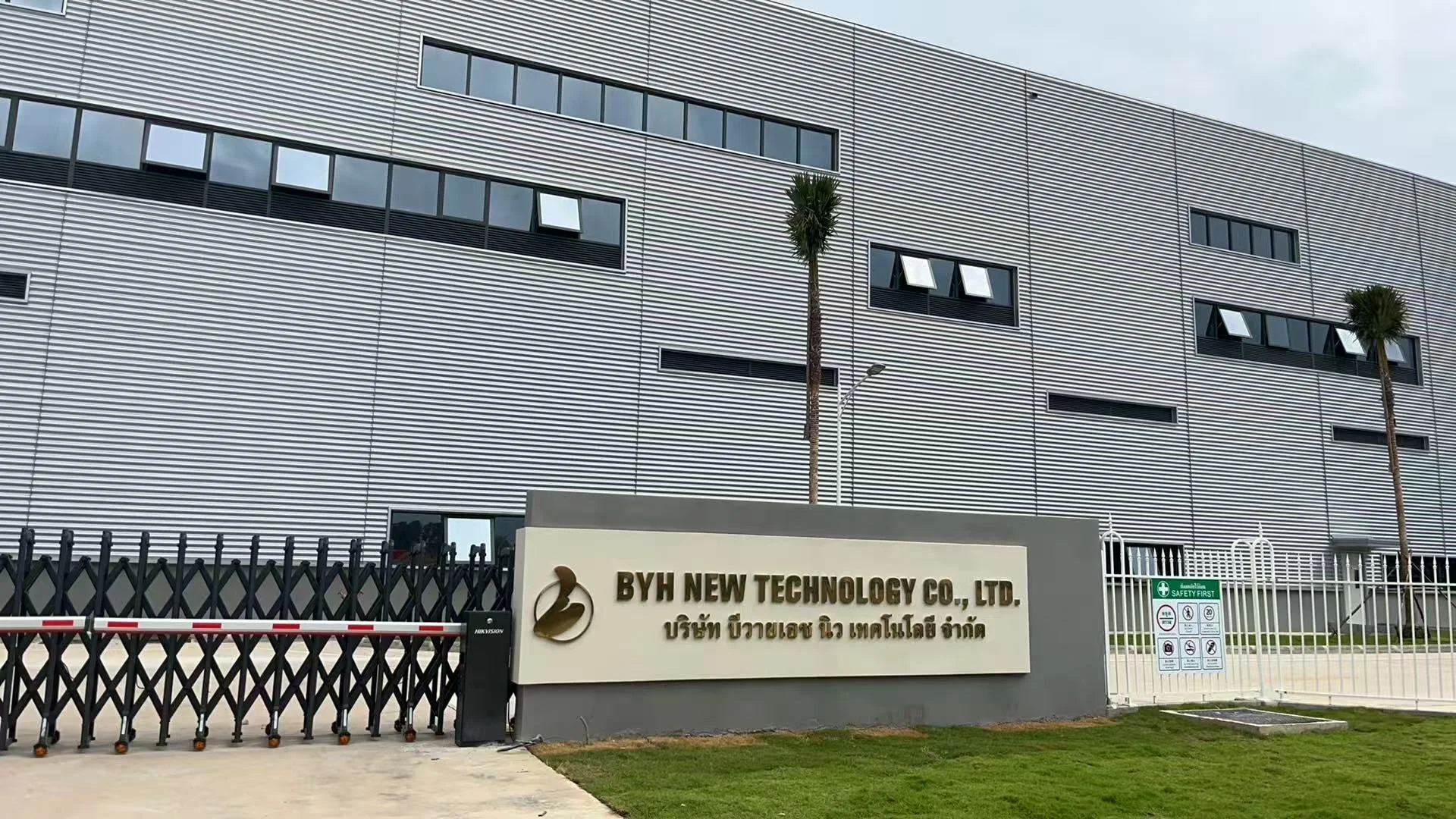Exterior of a modern industrial building with horizontal metal panel siding, surrounded by a black security gate, palm trees, and a sign displaying the name 'BYH New Technology Co., Ltd.' in both English and Thai, and safety instructions.