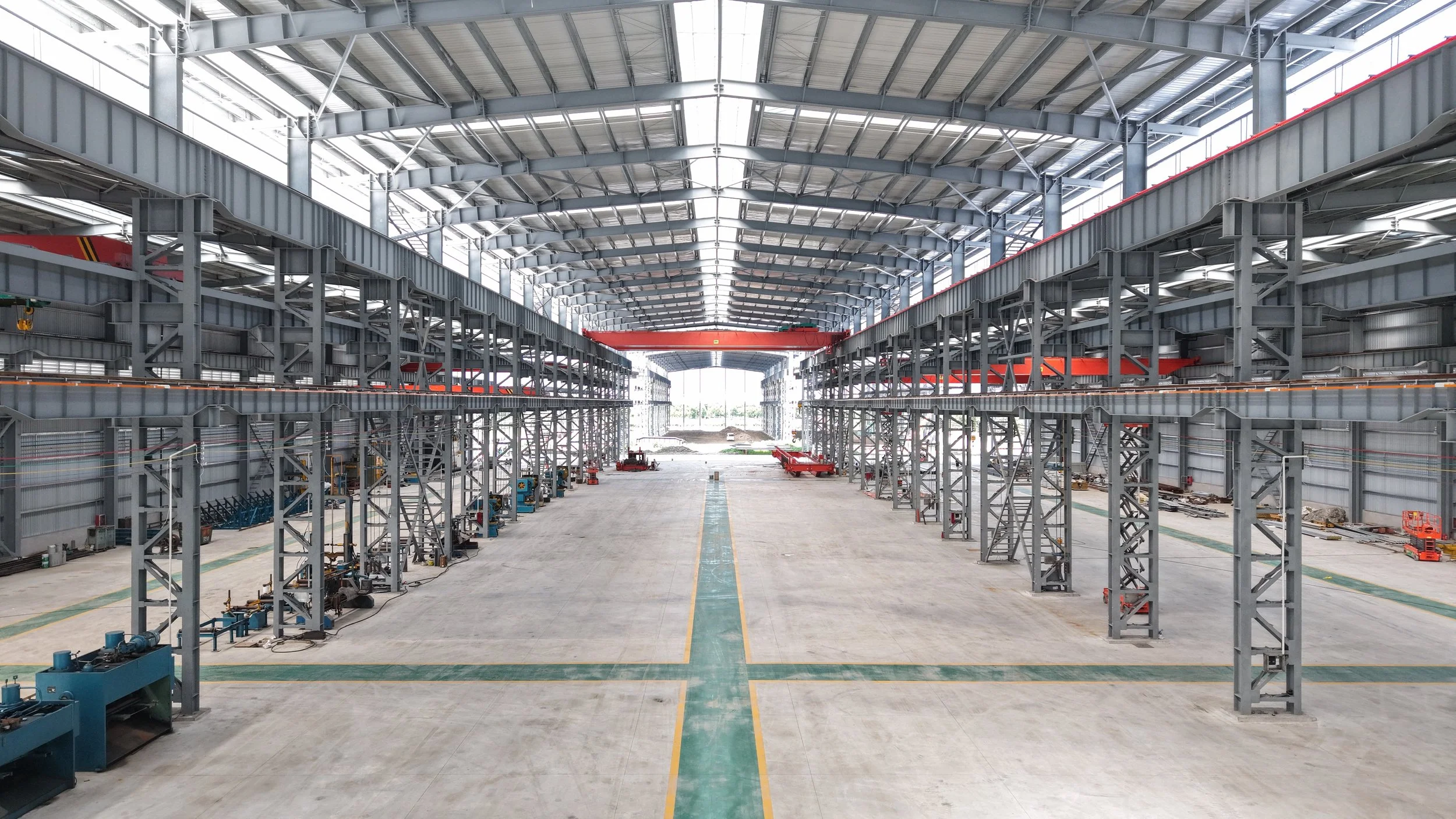 Interior of a large industrial warehouse with high ceilings, metal framework, and a wide open space with machinery along the sides.