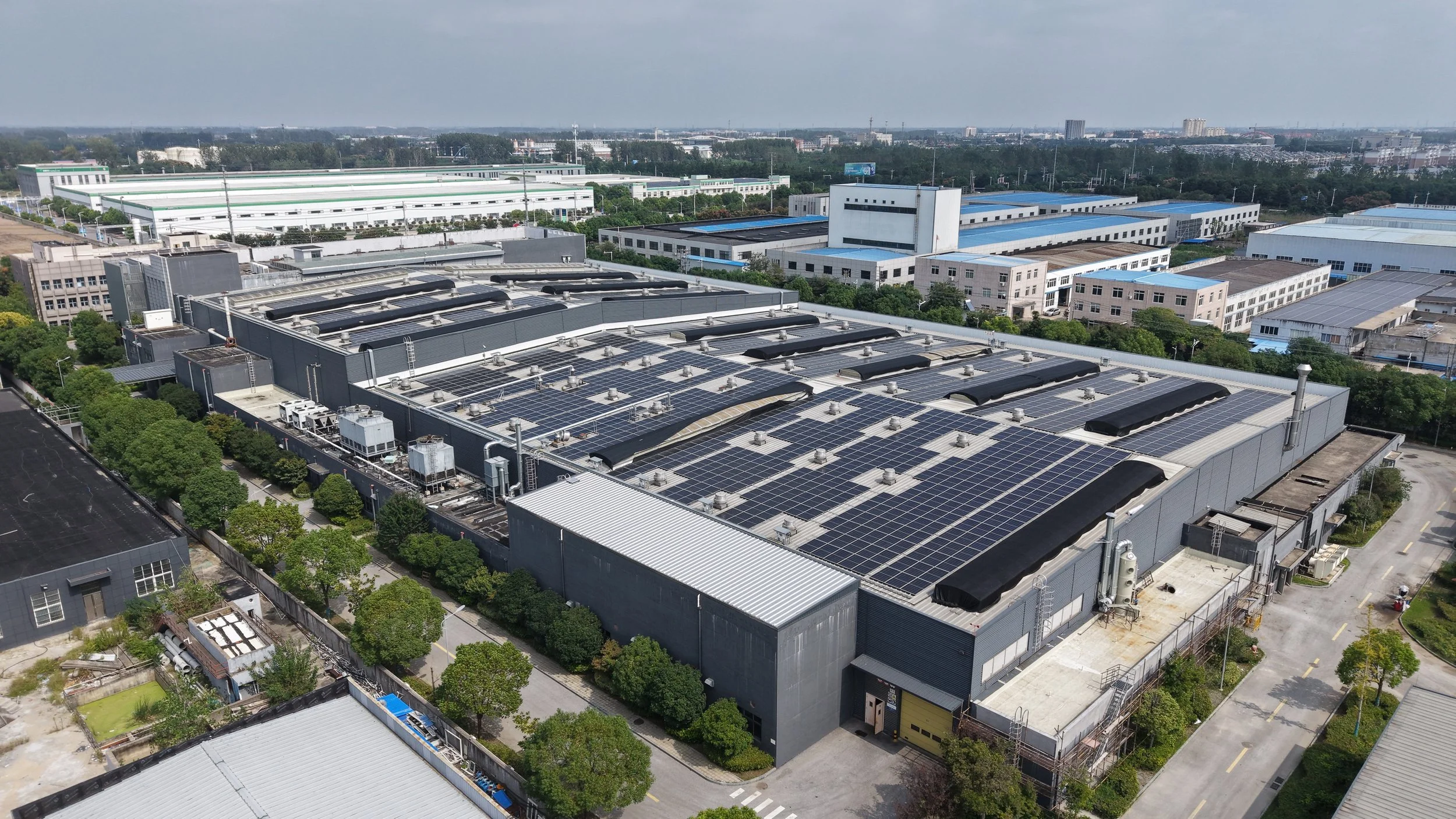 An aerial view of an industrial building with solar panels on its roof, surrounded by trees and other industrial buildings in the background.