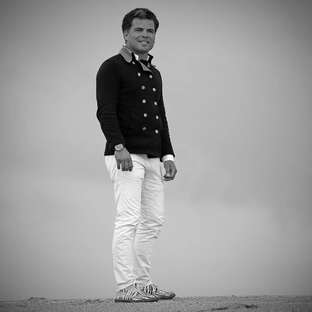 Monochrome image of a man wearing a double-breasted jacket, white pants, and patterned shoes, standing on a textured surface with a neutral background.