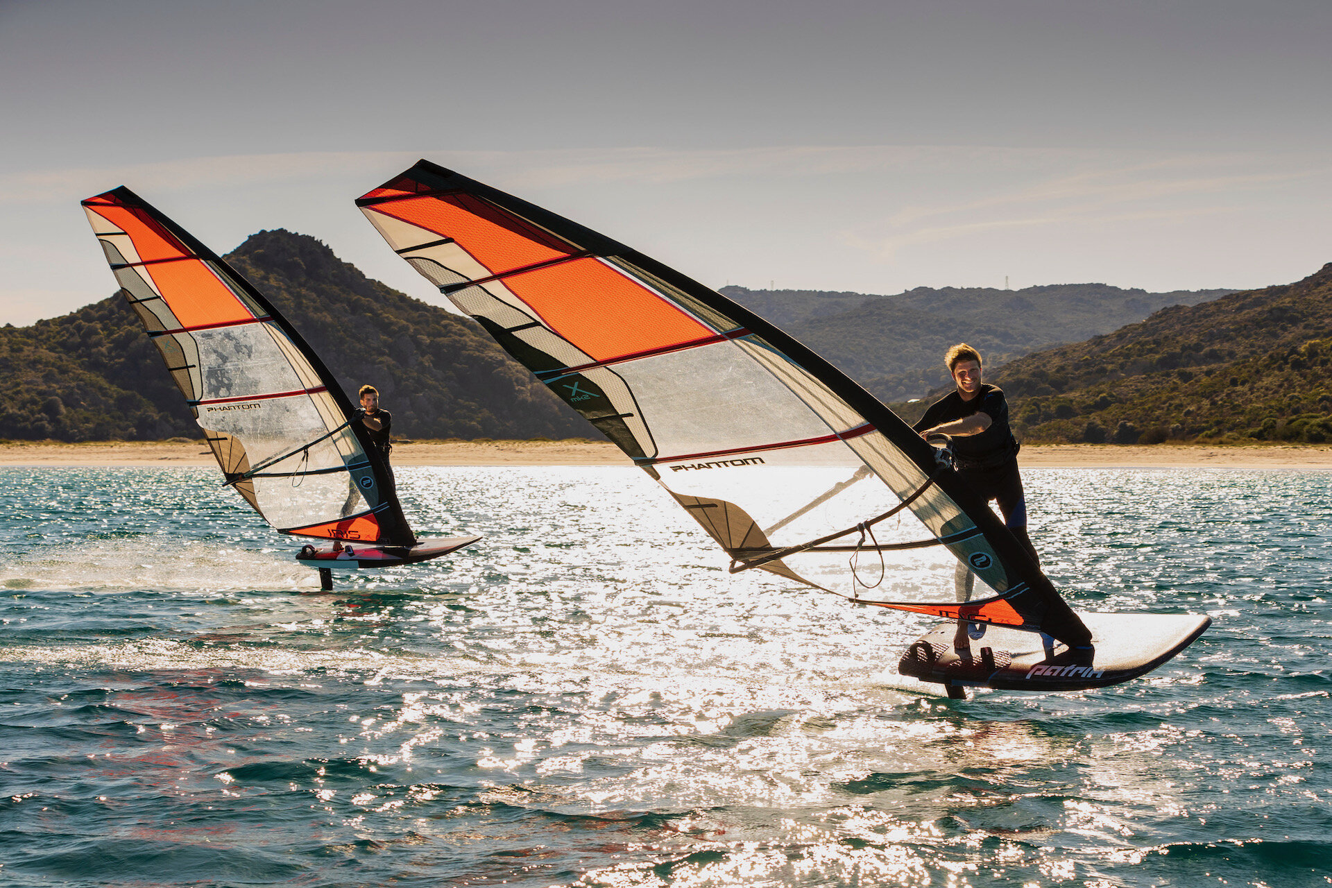 Two people windsurfing on clear blue water with mountainous landscape in the background.