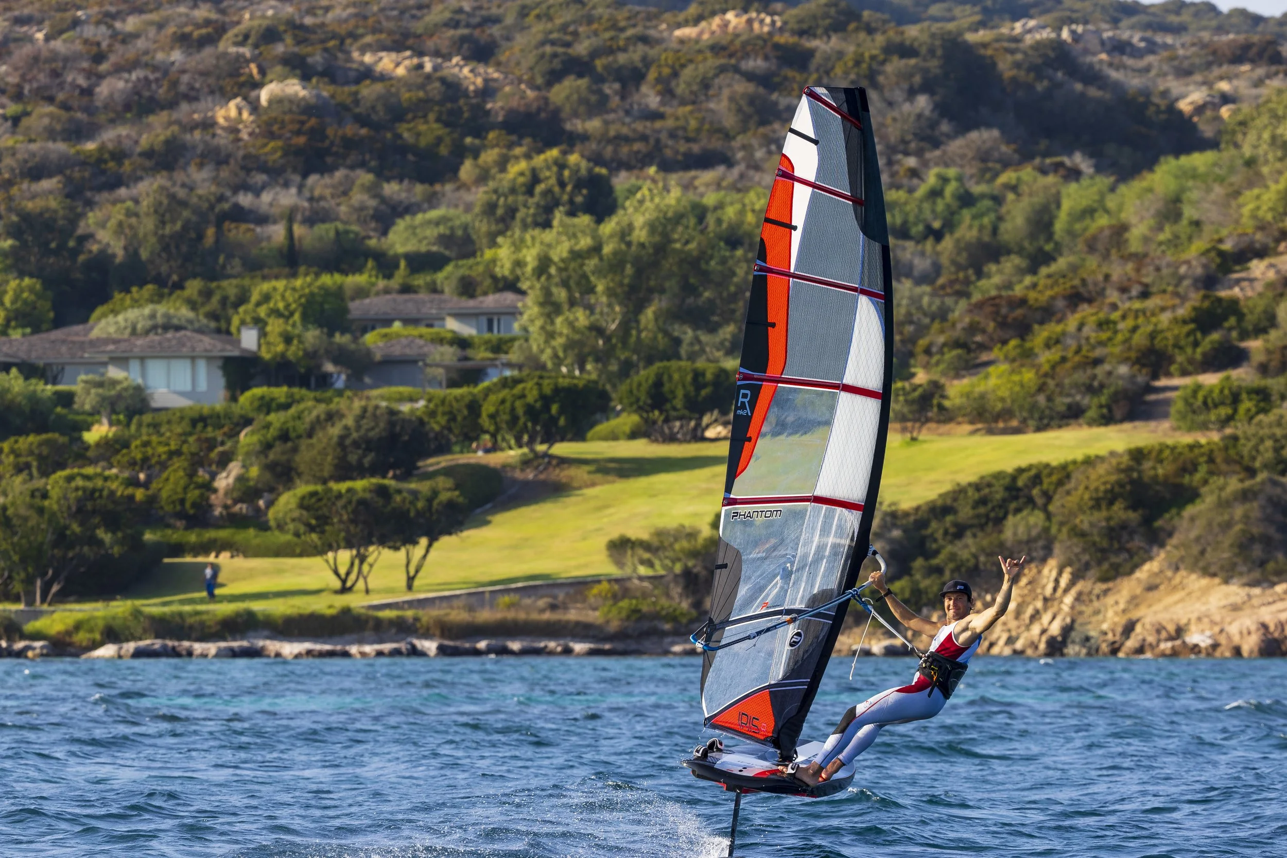 Person windsurfing on a hydrofoil board in front of a scenic, tree-covered hillside and houses.