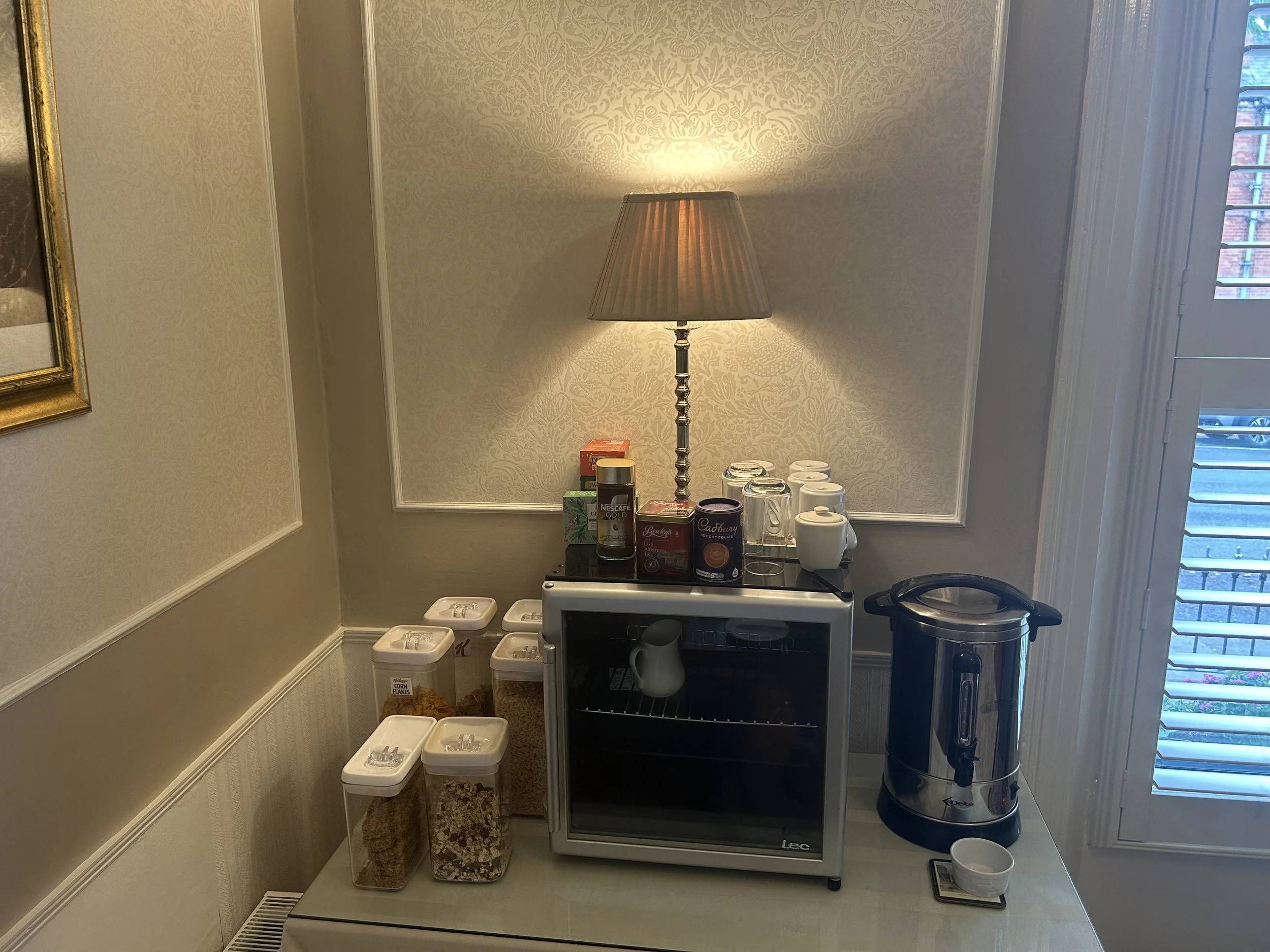Coffee station with jars of cereal, a small refrigerator, a lamp with a beige shade, glasses, coffee ingredients, and a tea/coffee maker on a white counter next to a window with shutters.