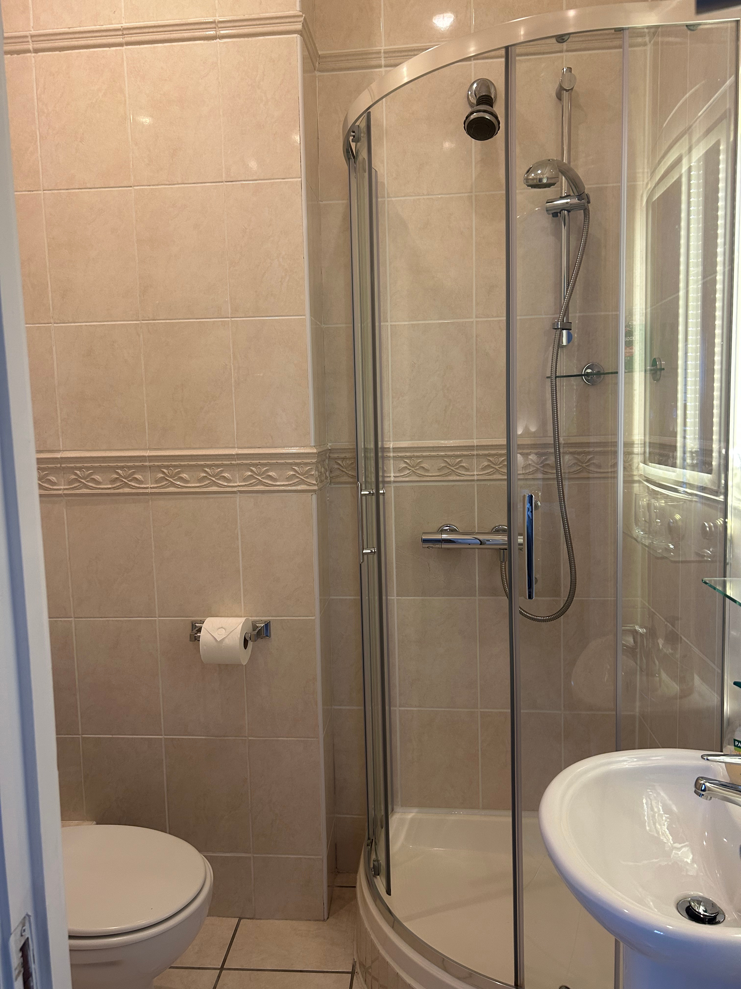 Small bathroom with beige tiled walls, a round corner shower with glass doors, a white pedestal sink, and a standard toilet with a toilet paper roll on the wall.