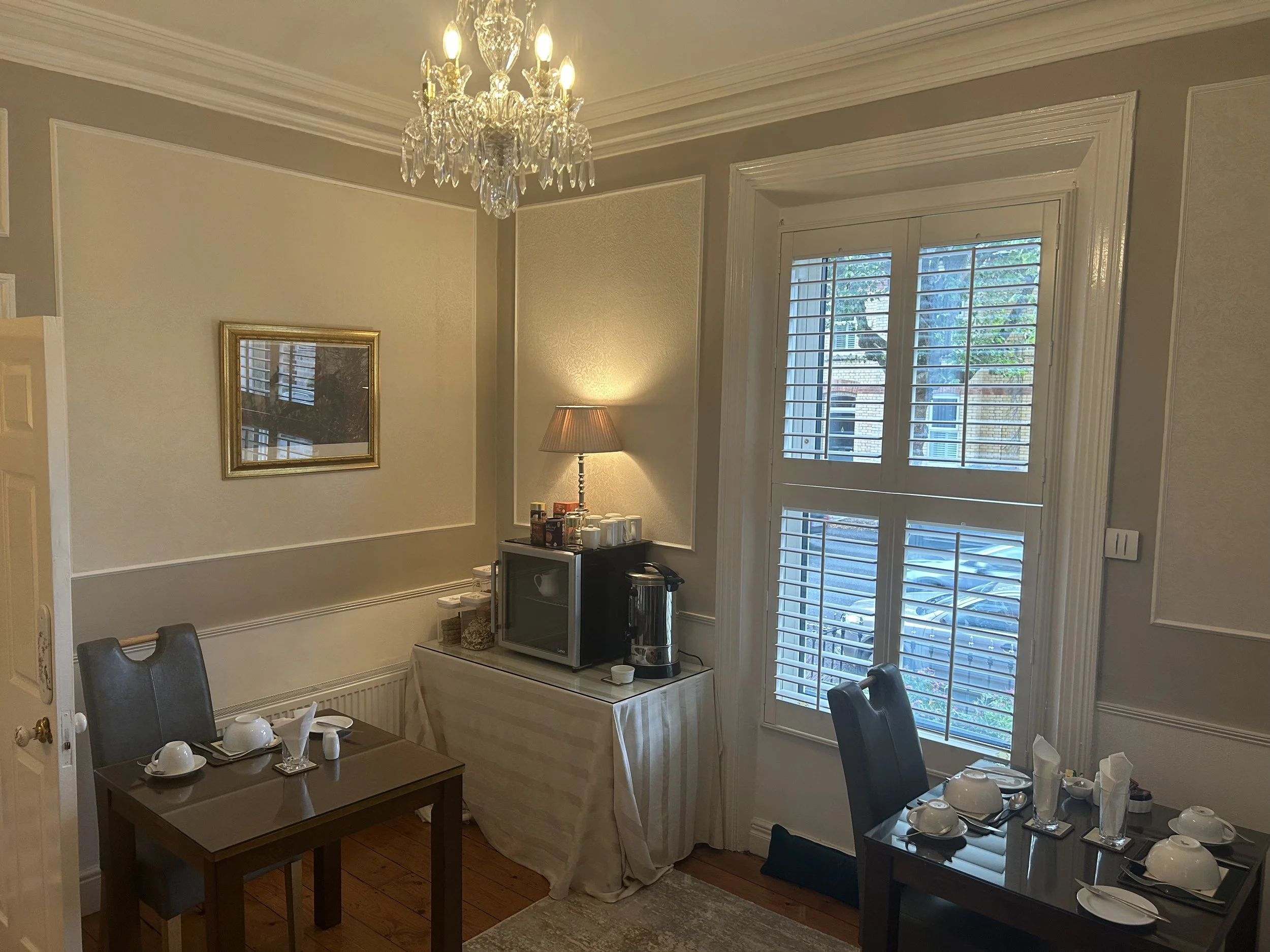 A dining room with a chandelier, two dark wood tables set for four, a side table with a microwave, coffee maker, and lamp, a window with white shutters, and framed artwork on the wall.