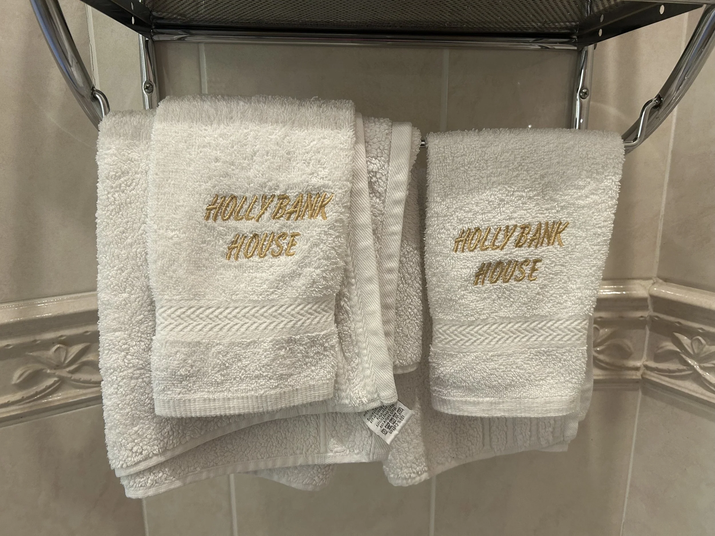 Two white embroidered towels with 'Hollybank House' on a bathroom shelf, one large and one small, against beige tiled wall.