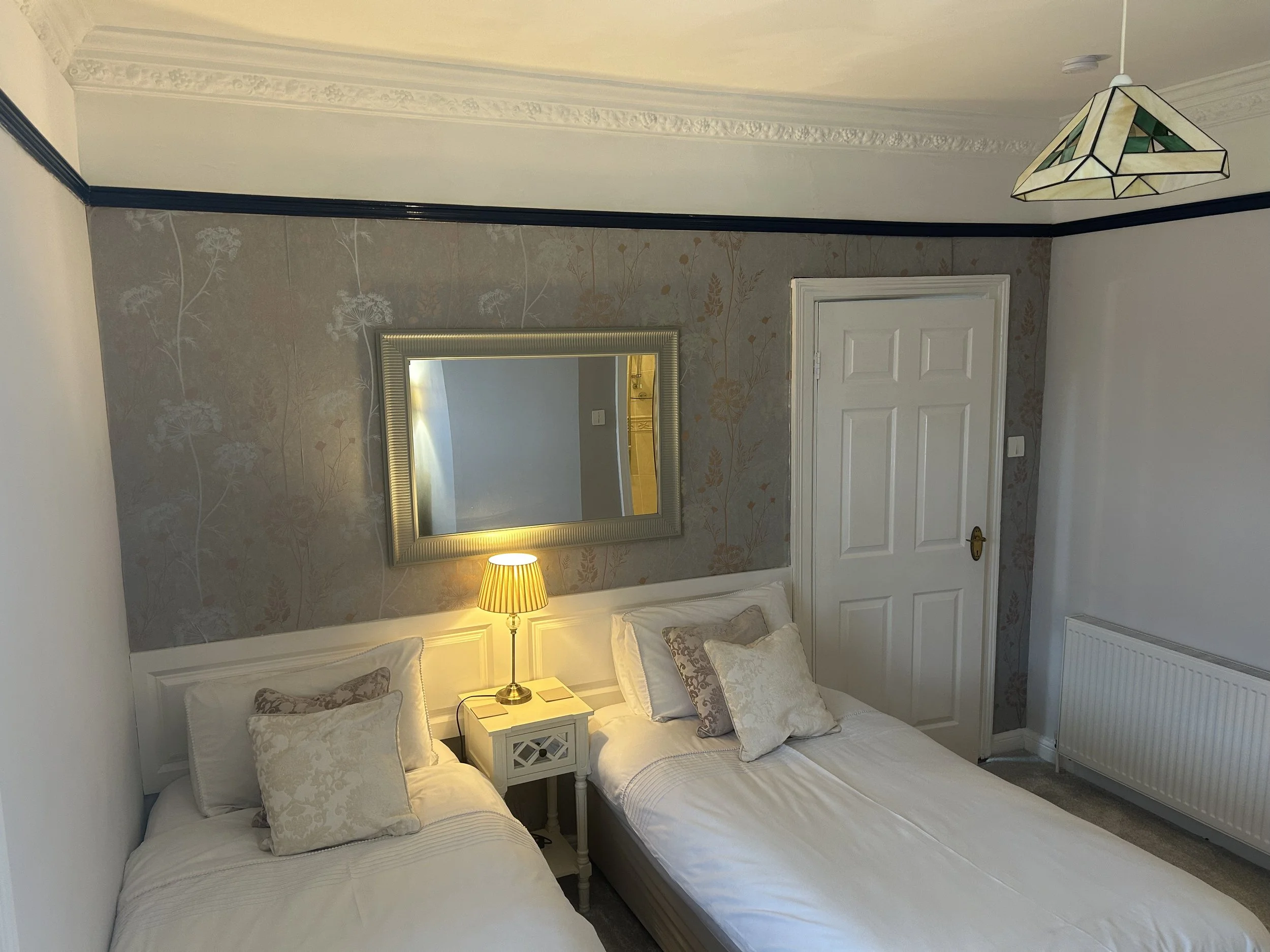A bedroom with two single beds, a bedside table with a lamp, a mirror above the beds, a closed white door, a radiator, and a stained-glass ceiling light. The wall behind the beds is decorated with patterned wallpaper.