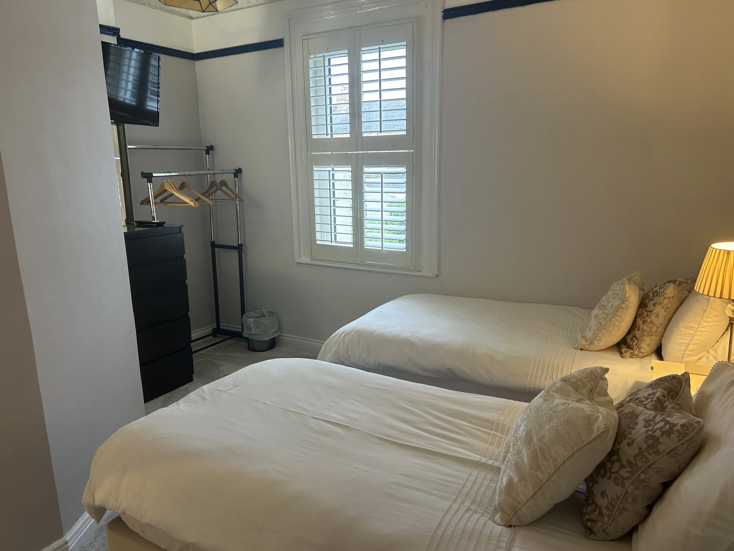 A bedroom with two twin beds covered in white bedding and pillows, a window with white shutters, a black dresser, a clothing rack with hangers, a small trash bin, and a lamp on a bedside table.