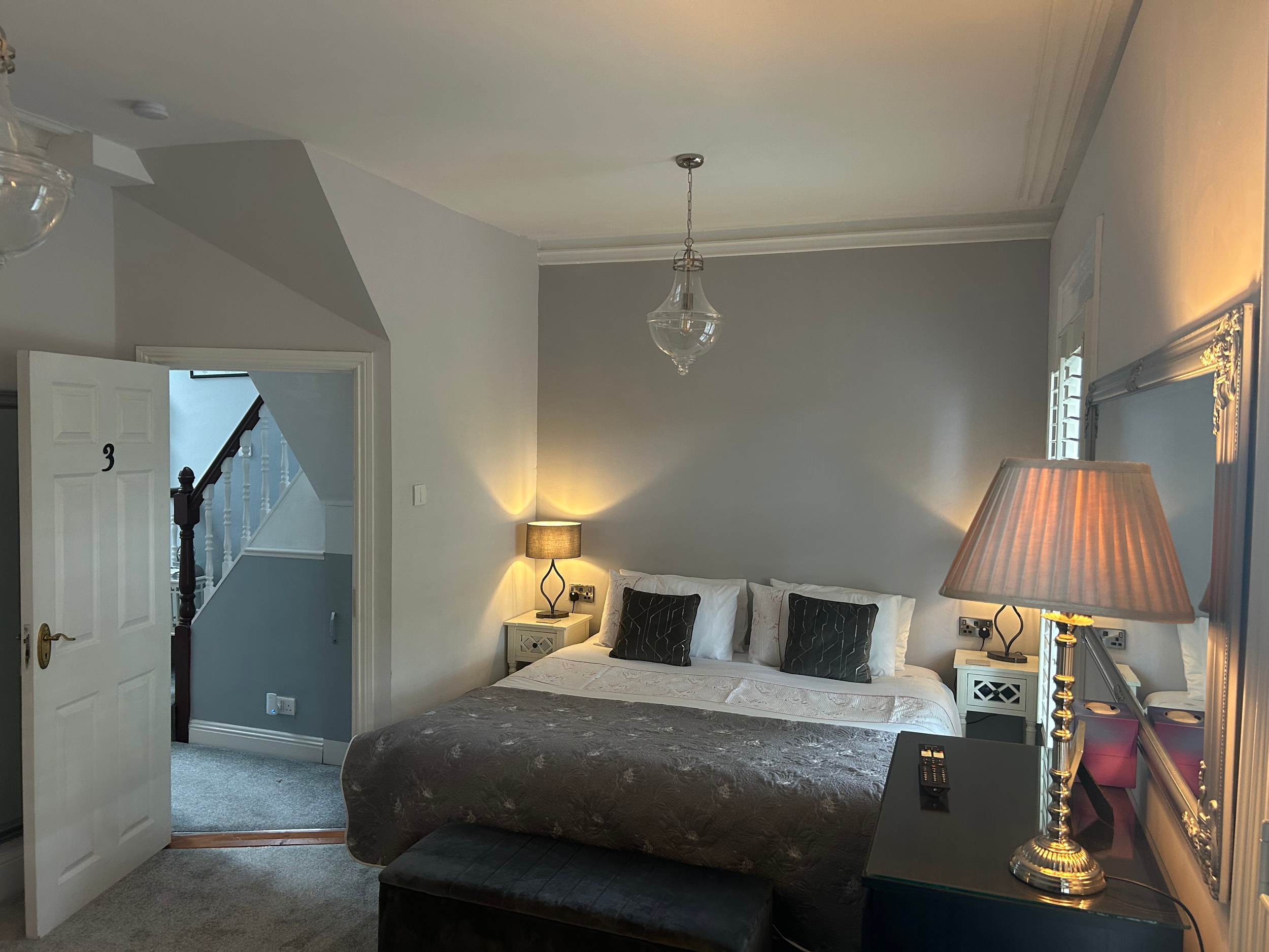 A cozy bedroom with a large bed, two nightstands with lamps, a mirror, and a view into a hallway with a staircase.