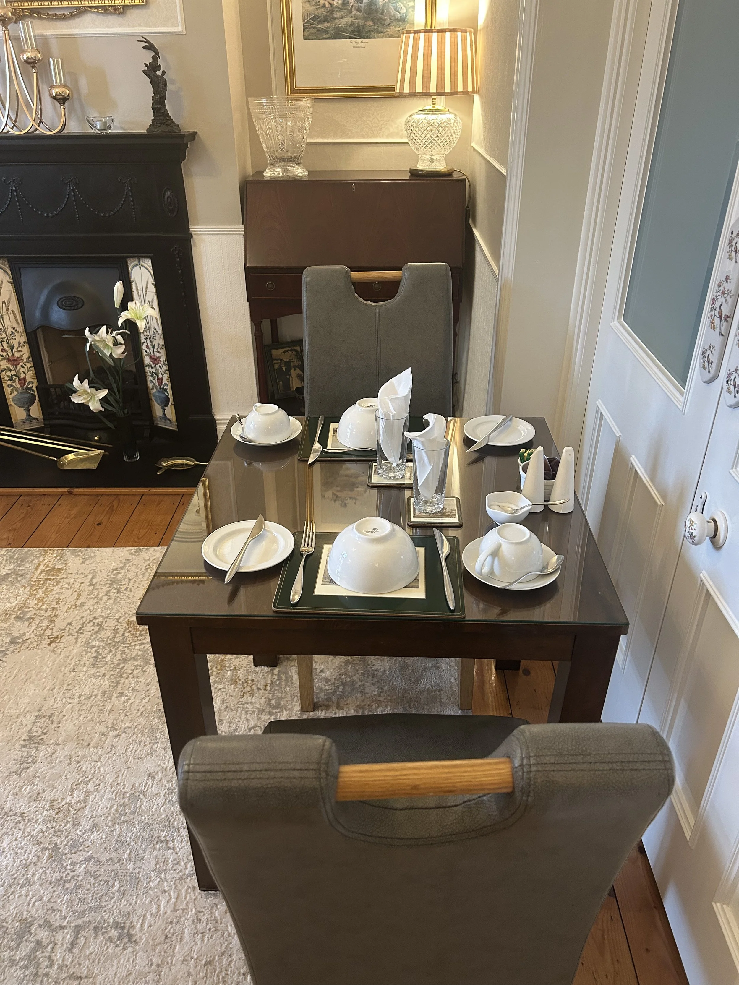 A dining table set for breakfast with white bowls, cups, and saucers, water glasses, napkins, and cutlery in a cozy, well-lit room with a fireplace, a sideboard, and decorative items.
