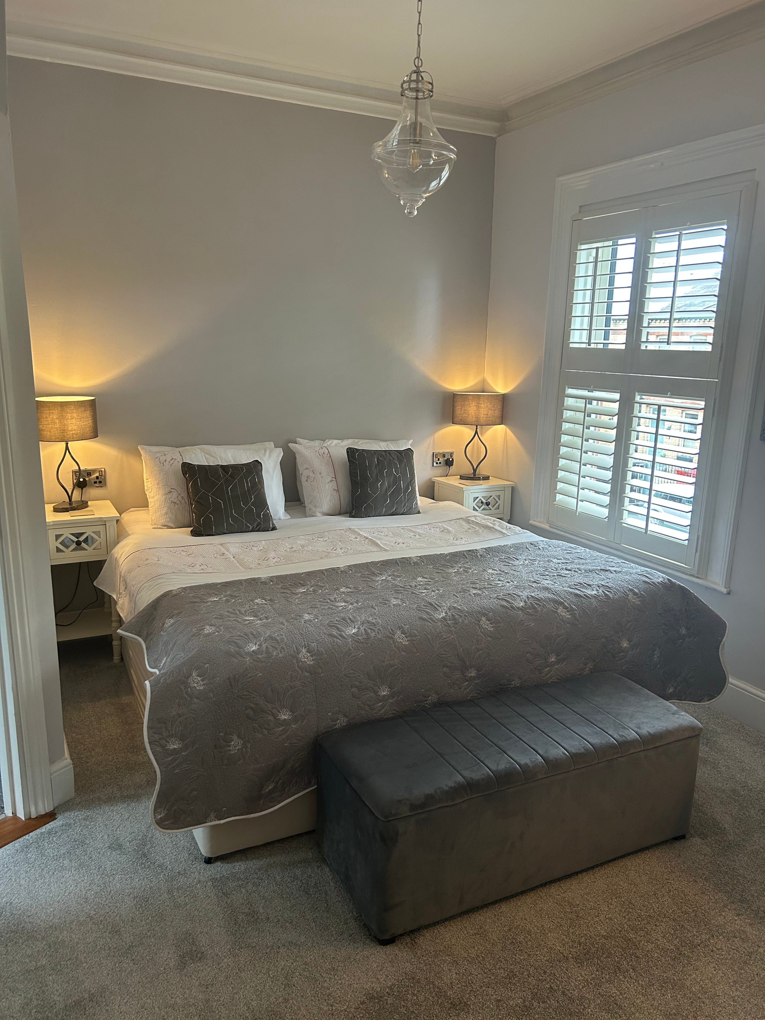 A bedroom with a large bed, two nightstands with lamps, a window with shutters, a gray upholstered bench at the foot of the bed, and a hanging glass light fixture.