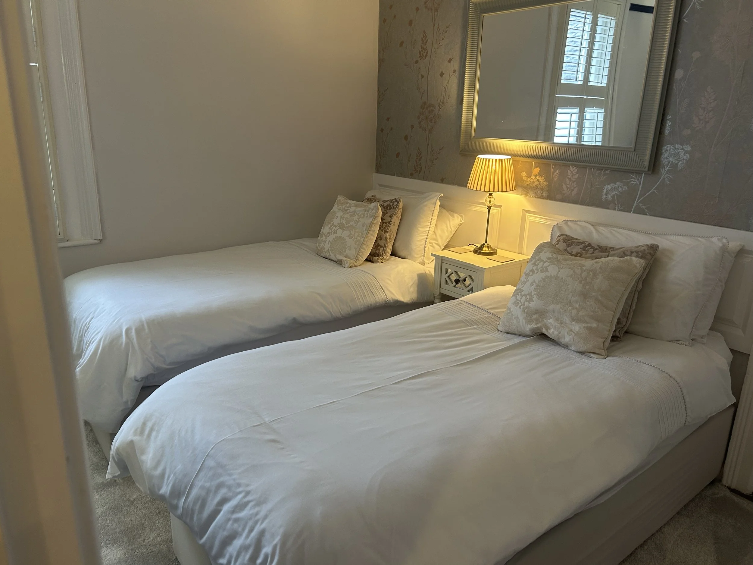 Two neatly made beds with white bedding and decorative pillows, a nightstand with a lampshade in a cozy bedroom, a rectangular mirror on a floral wallpapered wall, two windows with shutters