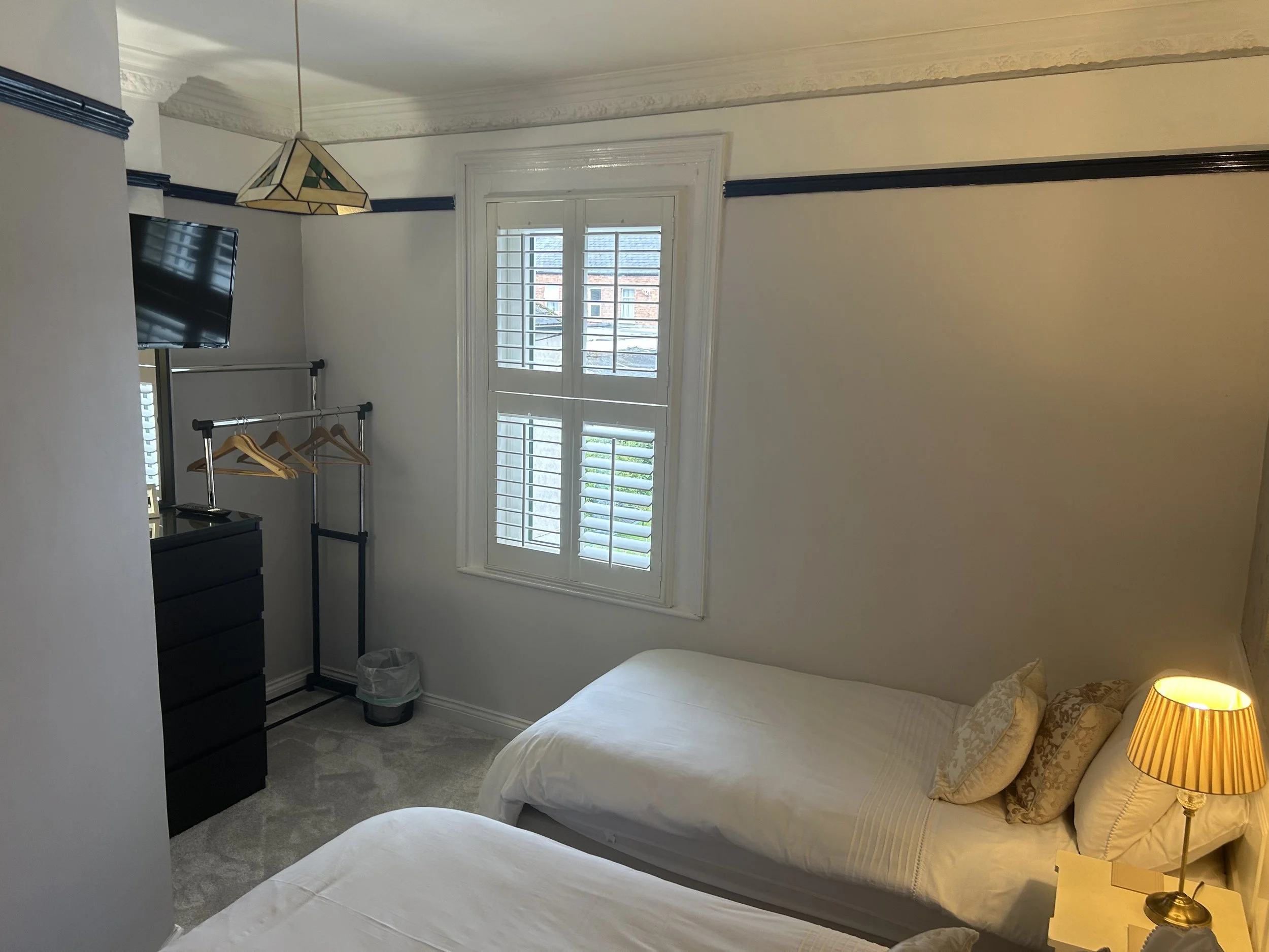 A small bedroom with two beds, white bedding, patterned pillows, a window with white shutters, a black dresser, a TV mounted on the wall, a clothing rack with wooden hangers, a small trash bin, and a lamp on a side table.