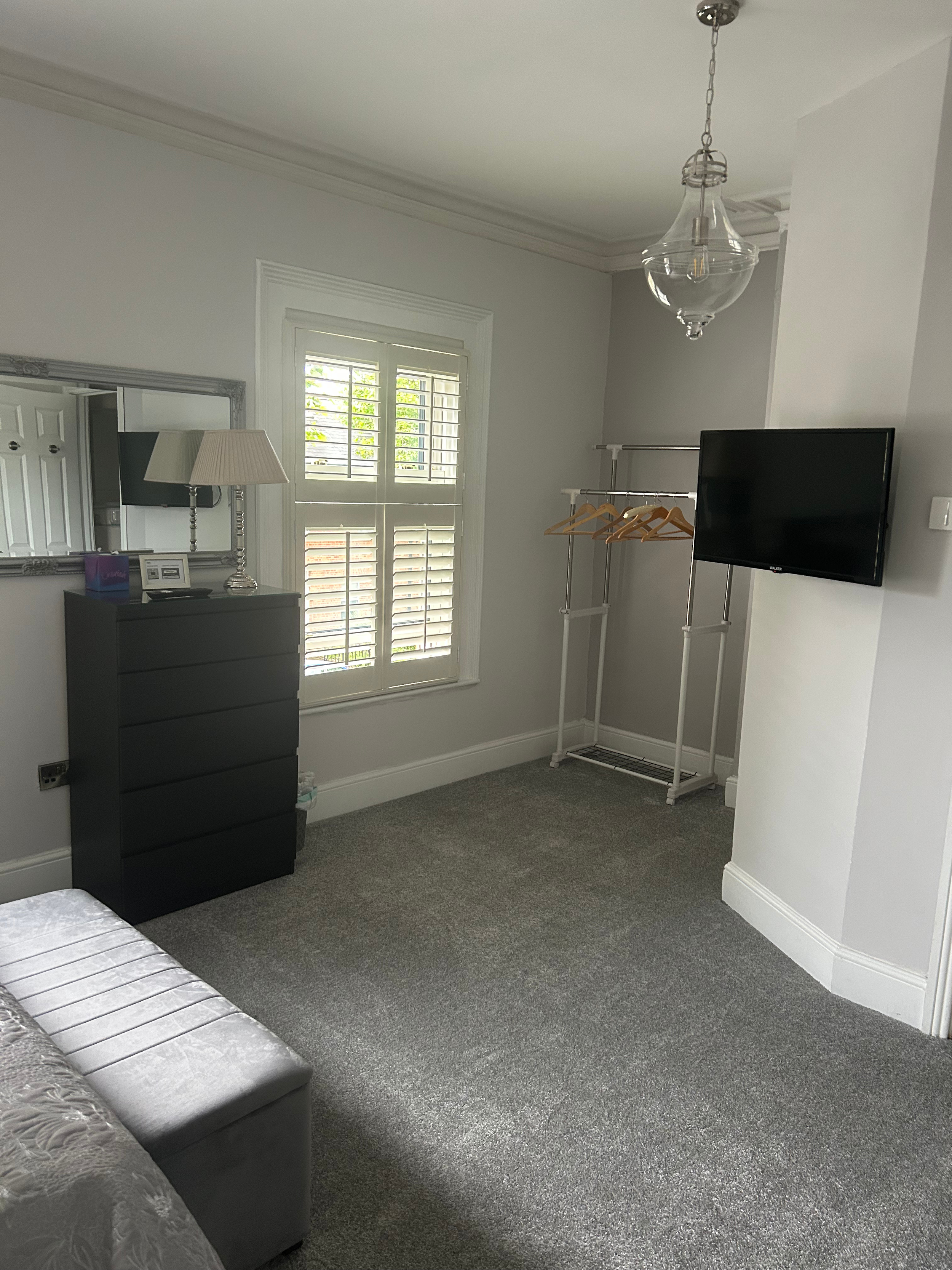 Empty bedroom with gray carpet, black dresser, mirror, window with plantation shutters, wall-mounted TV, and a clothing rack with hangers.