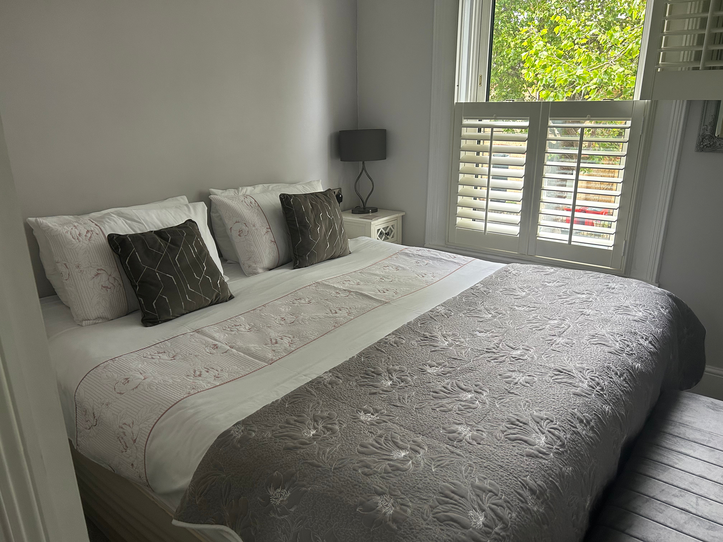 A neatly made bed with floral and dark accent pillows, a nightstand with a lamp, and a window with white shutters showing green trees outside.