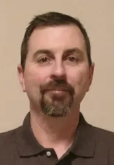 Portrait of a man with short dark hair, facial hair, and wearing a dark collared shirt, standing against a light-colored background.