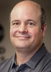 Close-up photo of a middle-aged man with a kind smile, wearing a dark shirt, in an indoor setting.