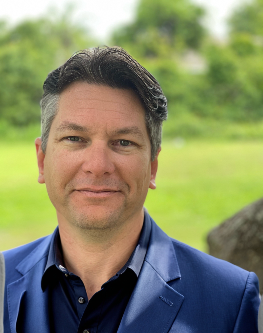 A man in a blue suit outdoors with greenery in the background.