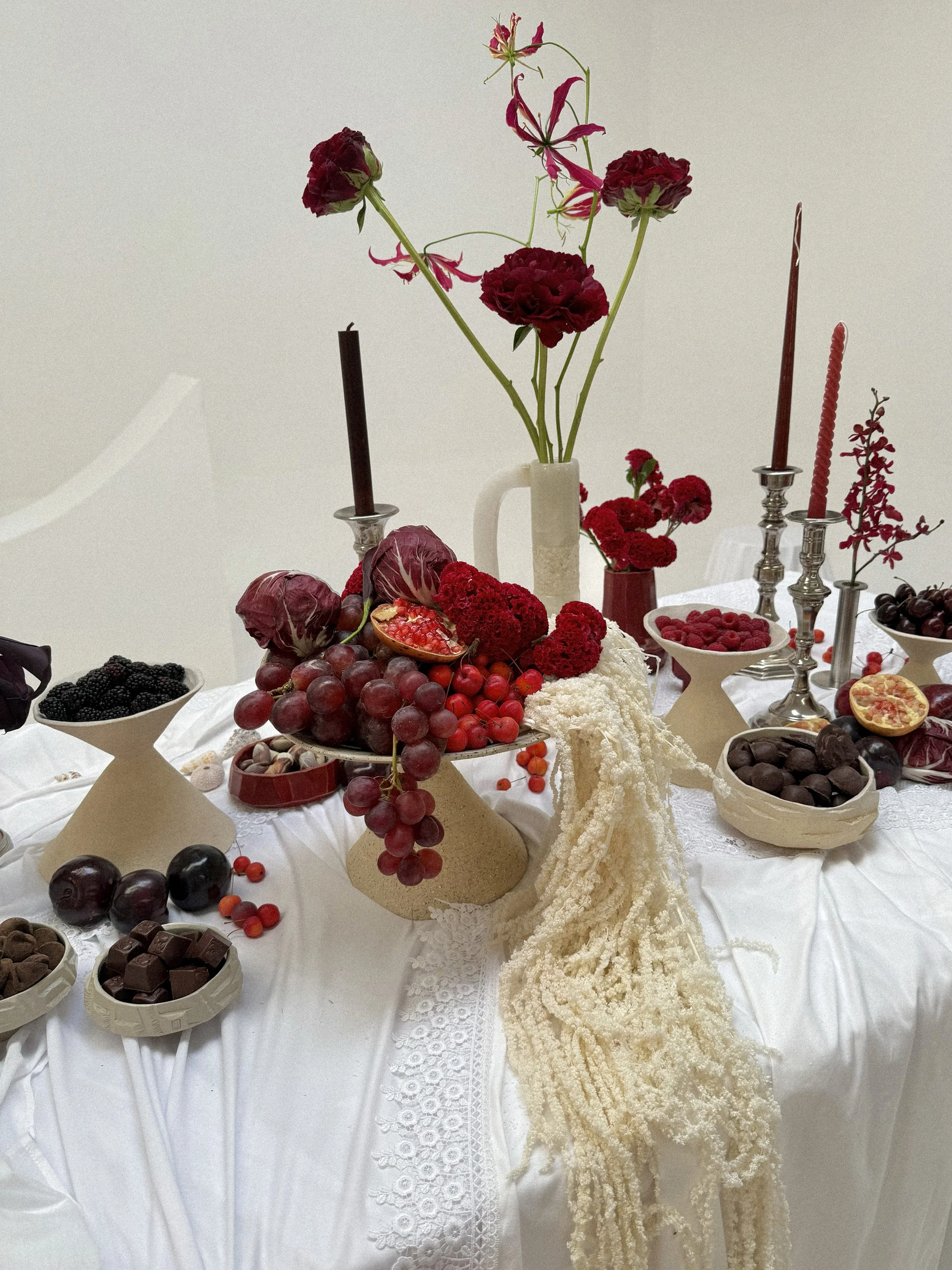 A table decorated with red and white flowers, candles, and a variety of fruits and chocolates, including grapes, blackberries, cherries, passion fruit, and assorted chocolates. The table has a white cloth with lace details.