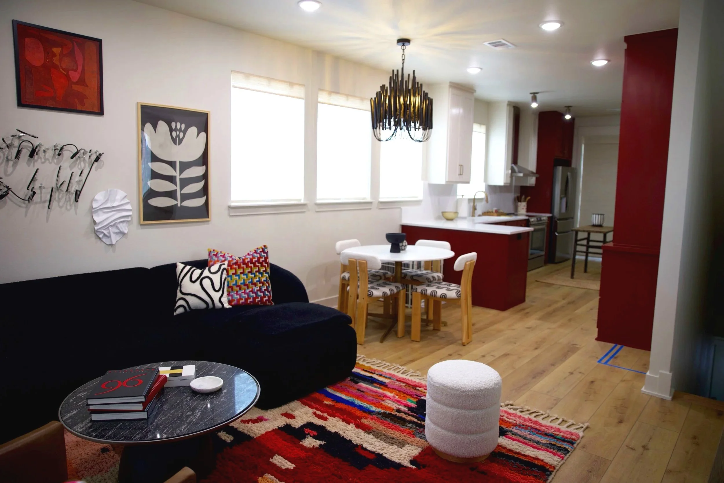 Living room and kitchen in an open-concept apartment. The living room features a black sofa with colorful throw pillows, a small round marble-topped coffee table with books and a dish, and a vibrant striped rug. The wall has abstract art and metal wa