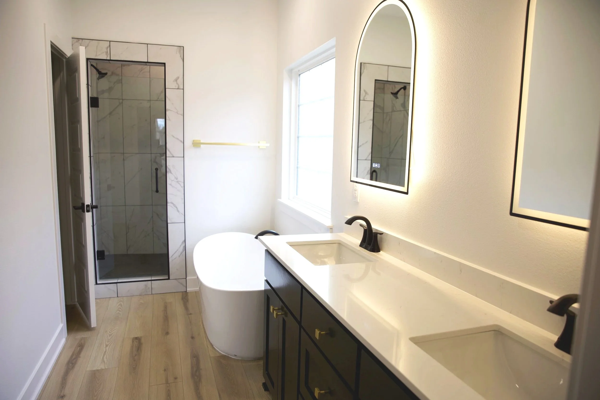 Modern bathroom with a walk-in shower, double vanity with black fixtures, and a freestanding bathtub. Light-colored walls and wooden flooring.