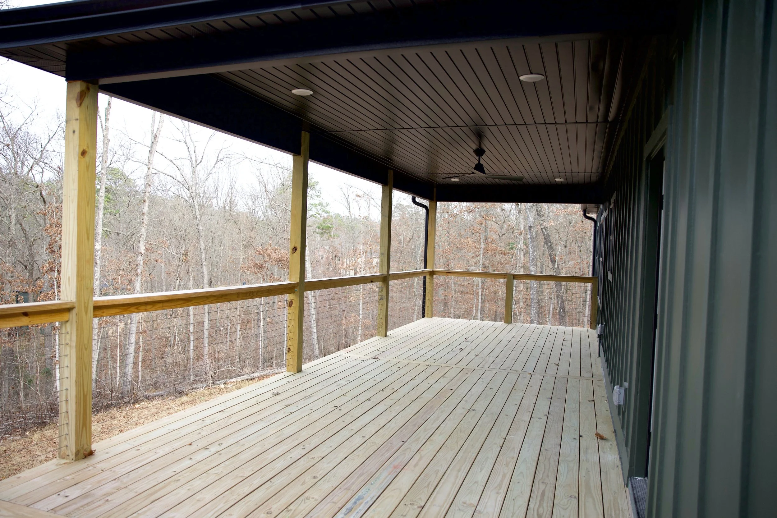 A spacious outdoor wooden deck with railings, attached to a green building, overlooking a wooded area with leafless trees.