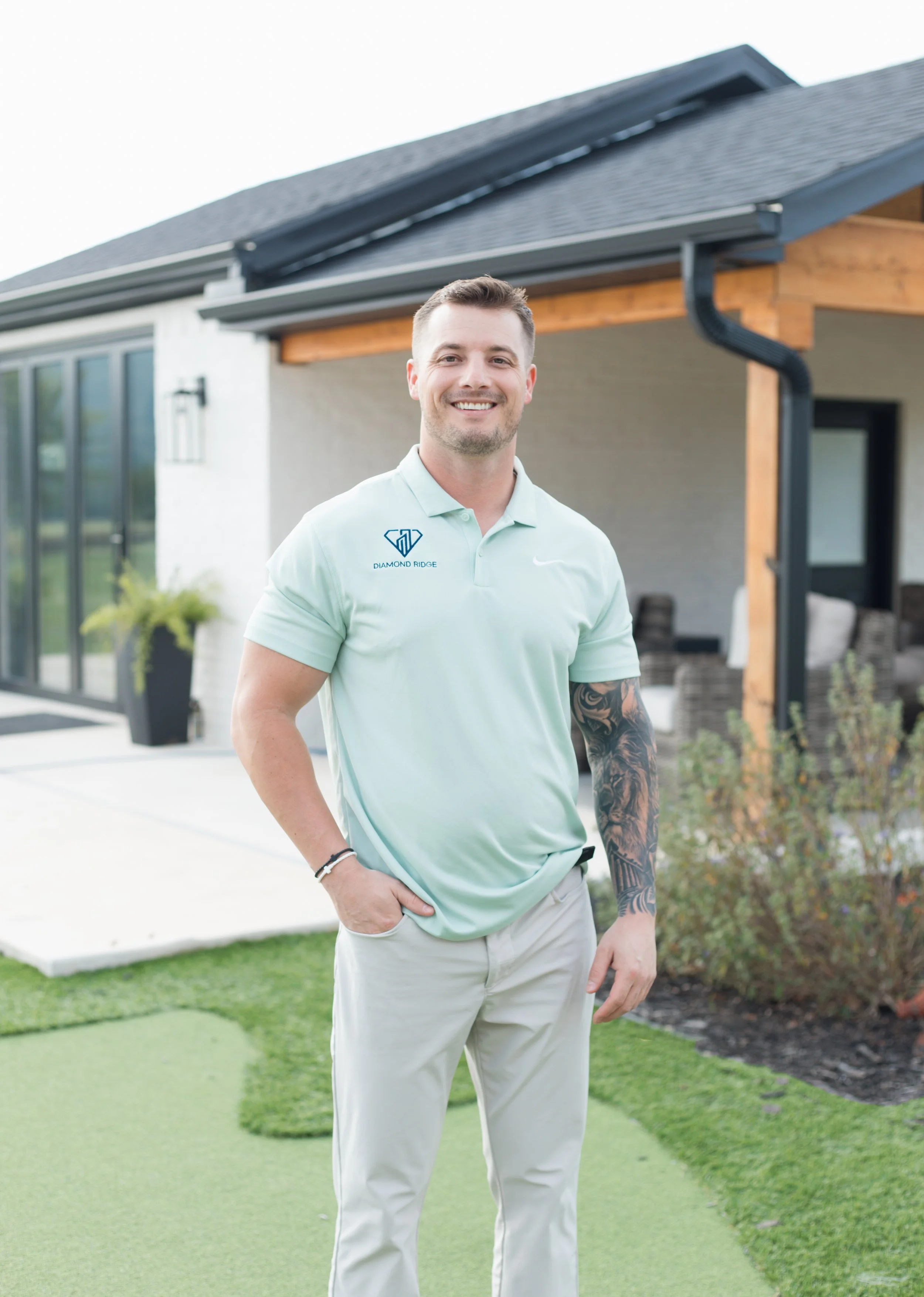 A smiling man standing outdoors in front of a modern house with a porch, wearing a light green polo shirt with an embroidered logo on the chest and light-colored pants.