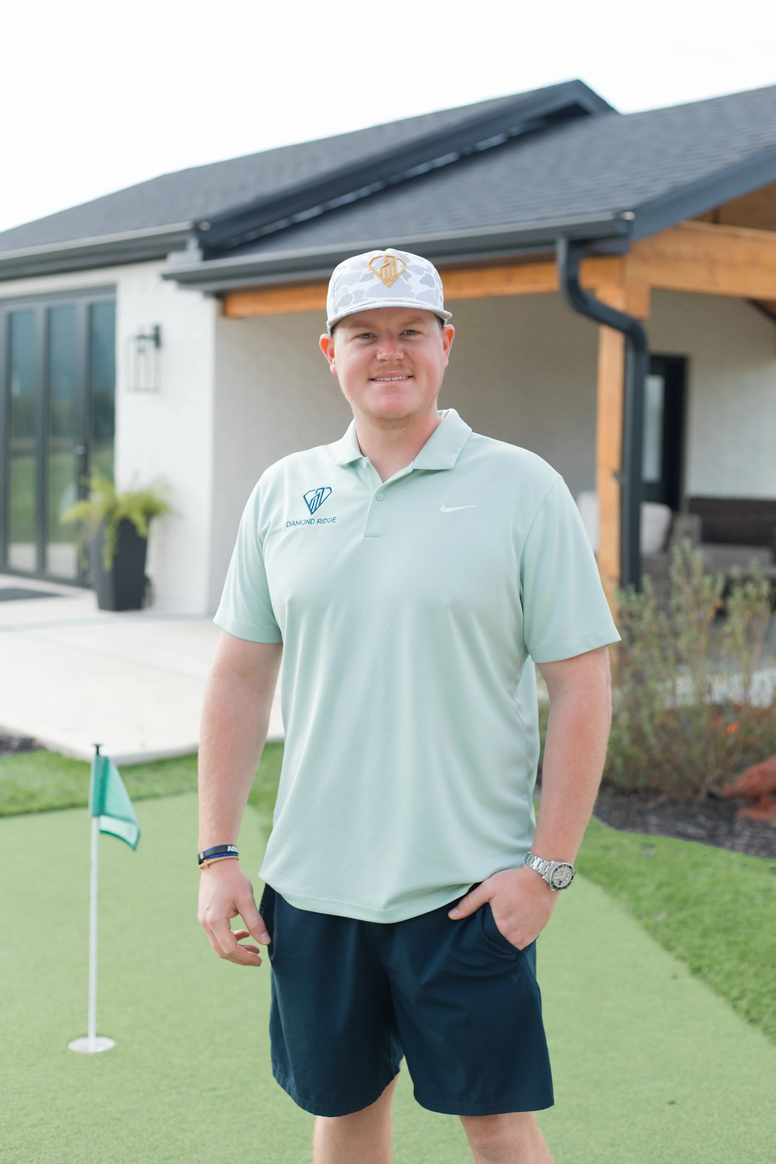 A man standing on a golf course near the putting green, wearing a light-colored golf shirt with a logo and the words "Diamond Ridge," navy shorts, a watch, bracelets, and a baseball cap, with a house and garden in the background.