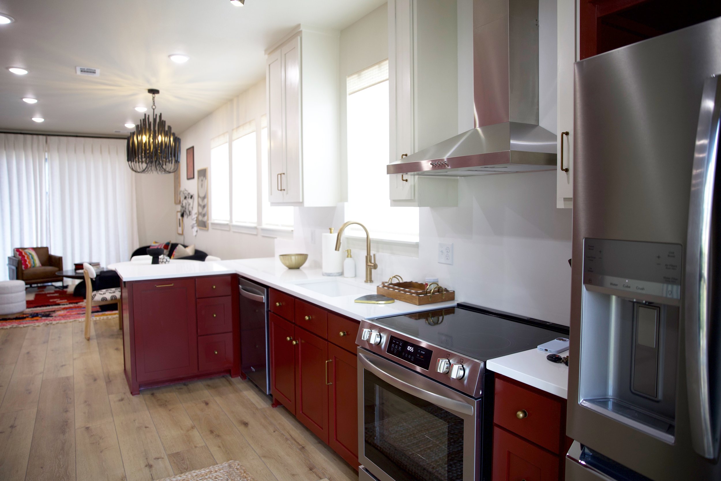 Open kitchen with red cabinets, white countertops, stainless steel oven, and refrigerator, overlooking a living area with armchairs and a chandelier.