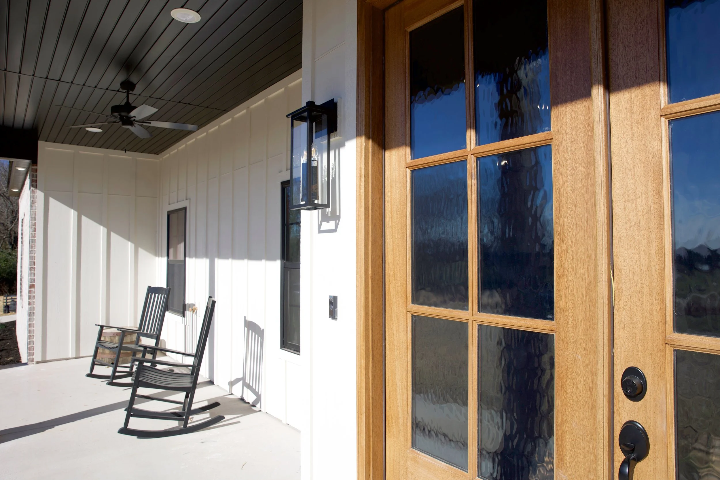 Front porch of a house with two black wooden rocking chairs, a black doorbell, a black outdoor wall lantern, and a wooden-framed glass door. The porch has a ceiling fan and a modern light fixture, with sunlight casting shadows on the white wall.