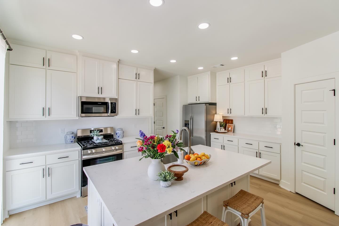 Bright white kitchen with central island, stainless steel appliances, including refrigerator, microwave, and oven; white cabinetry; decorative plants and flowers on the island and counter; bowl of peaches; small table lamp with a framed photo nearby.