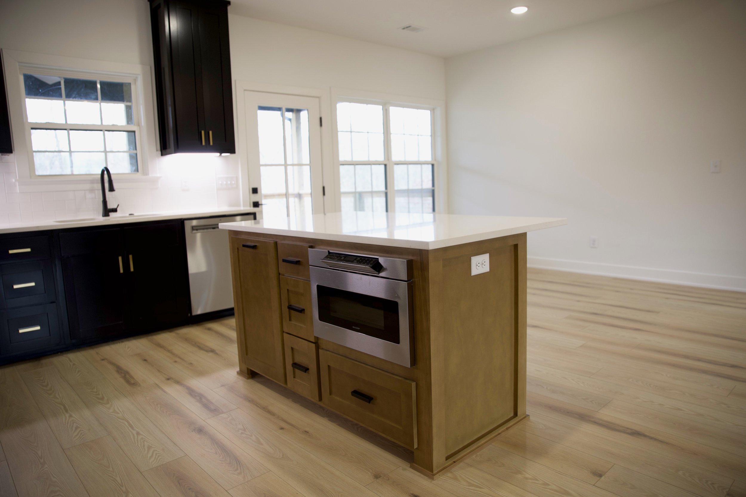 Modern kitchen with wood flooring, an island with a built-in oven, black and white cabinets, a sink, large windows, and a bright open space