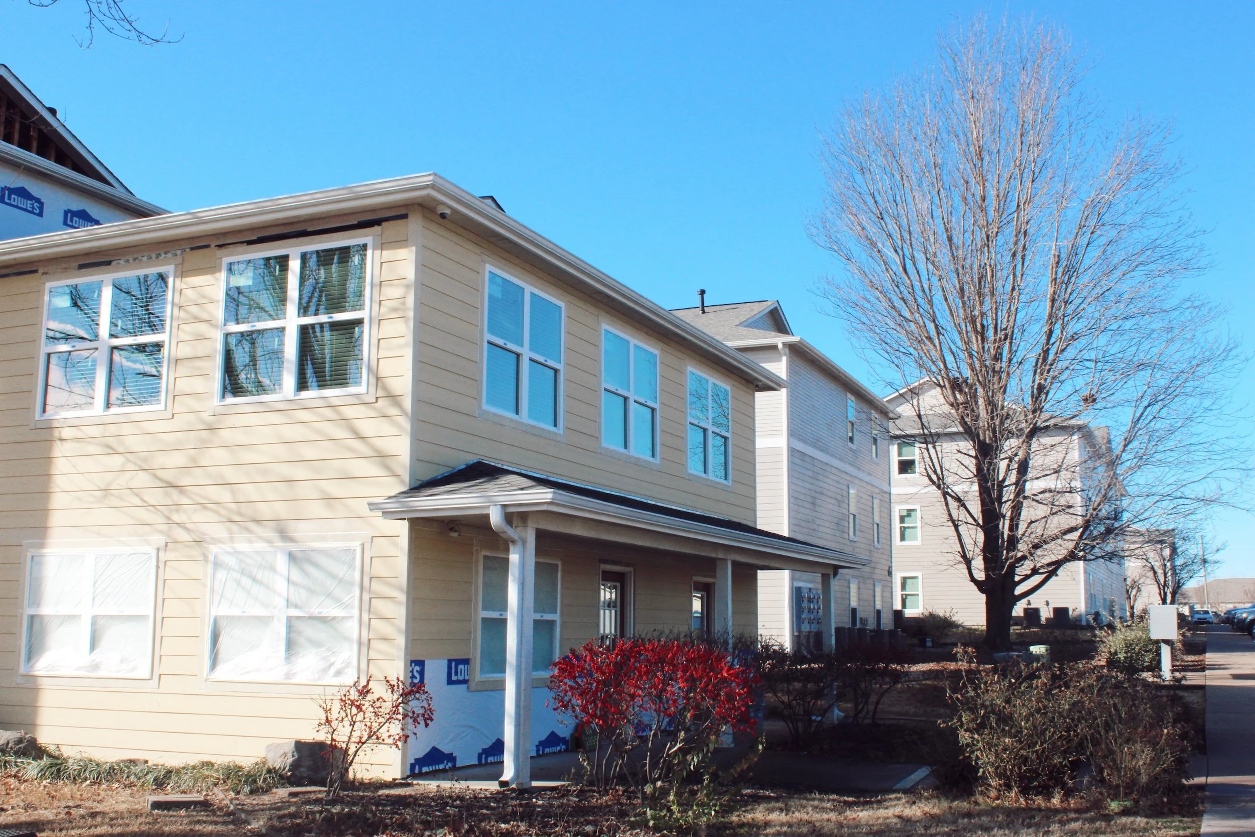 A newly built beige and white multi-family residential building with light-colored siding, multiple windows, small porch, and a large leafless tree in front, under clear blue sky.