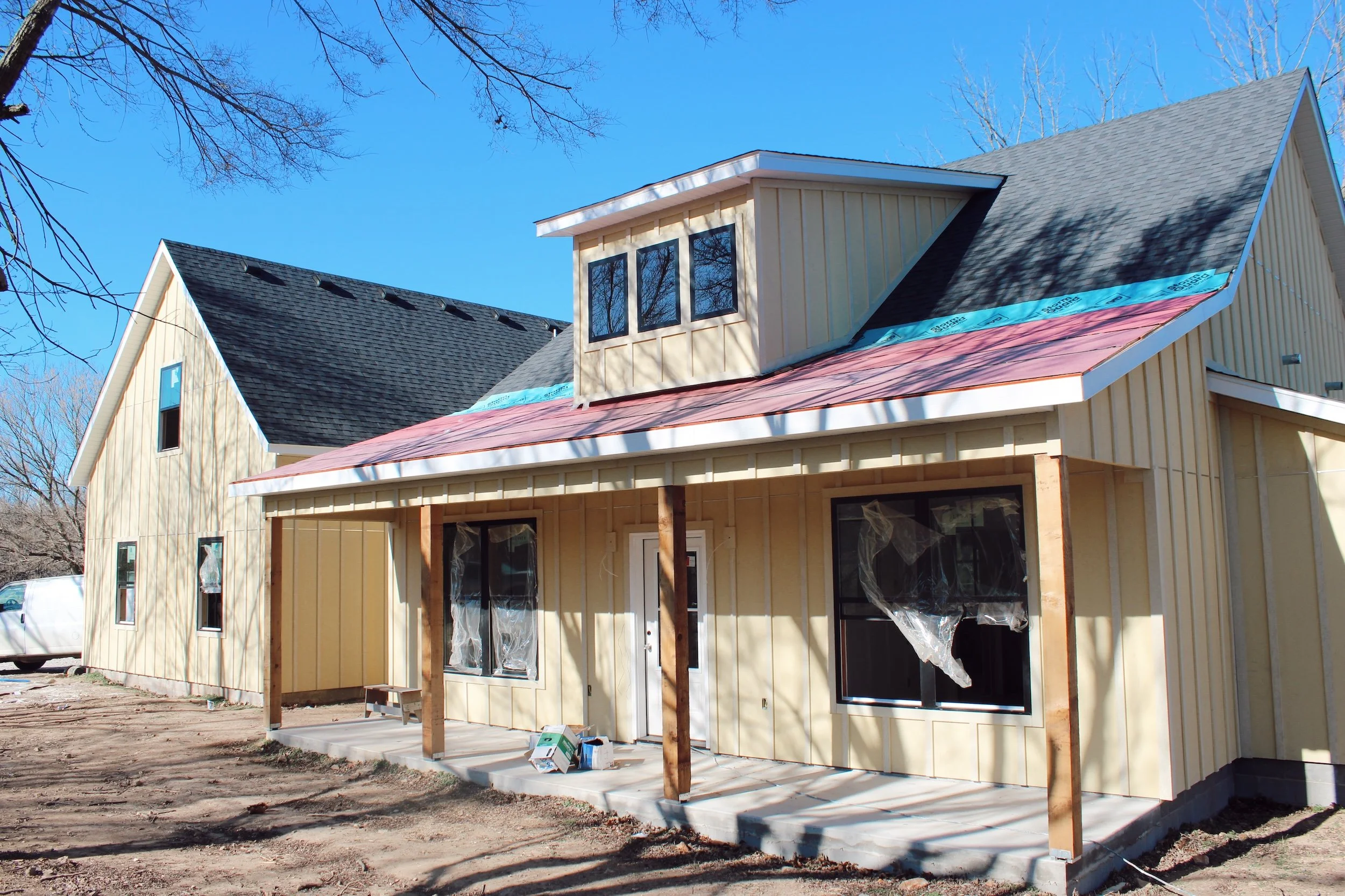 A house under construction with beige vertical siding, an unfinished front porch, and a partially installed metal roof, set against a clear blue sky.