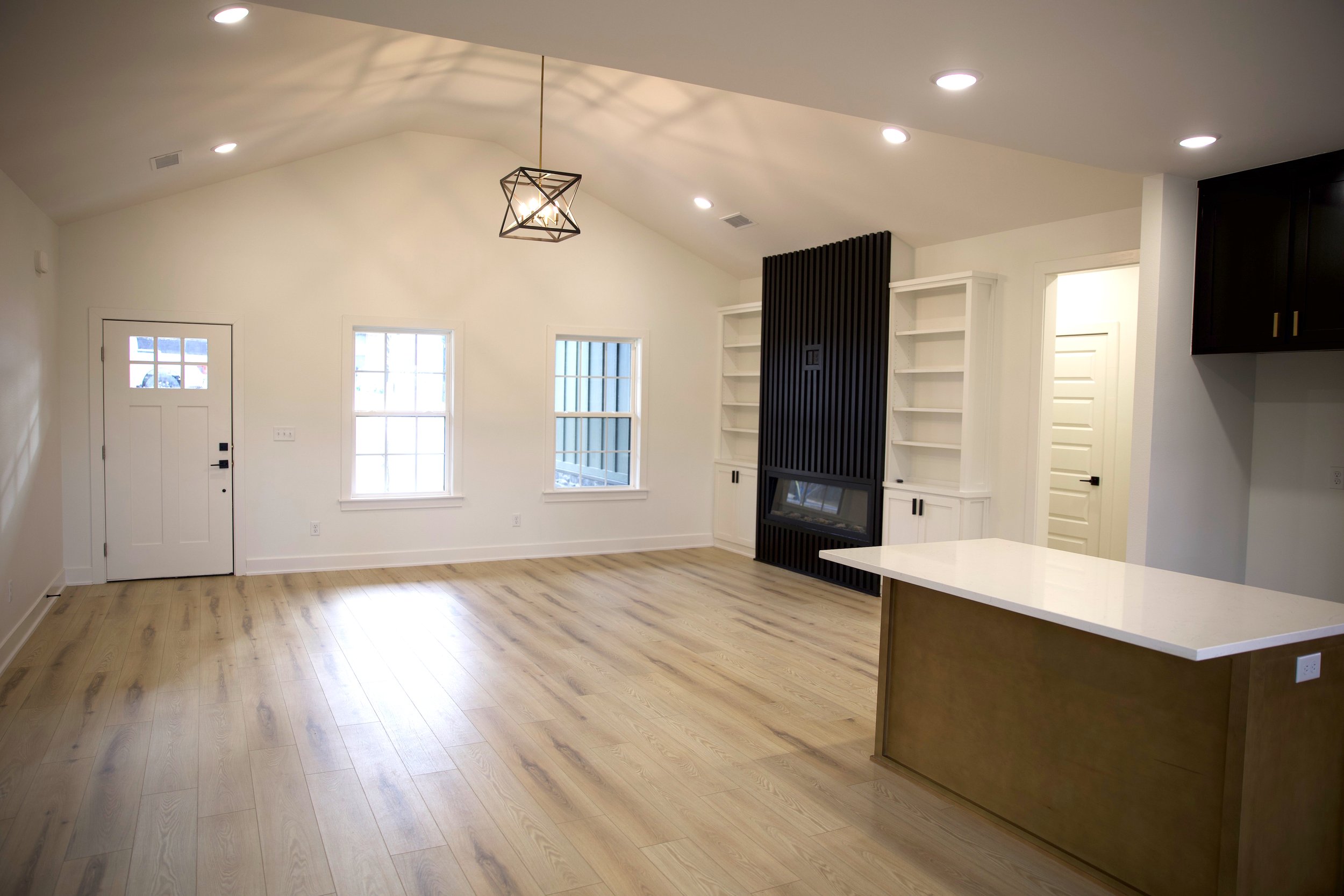 Empty living room with white walls, large windows, a black fireplace with white shelves, wooden flooring, and a white countertop island.